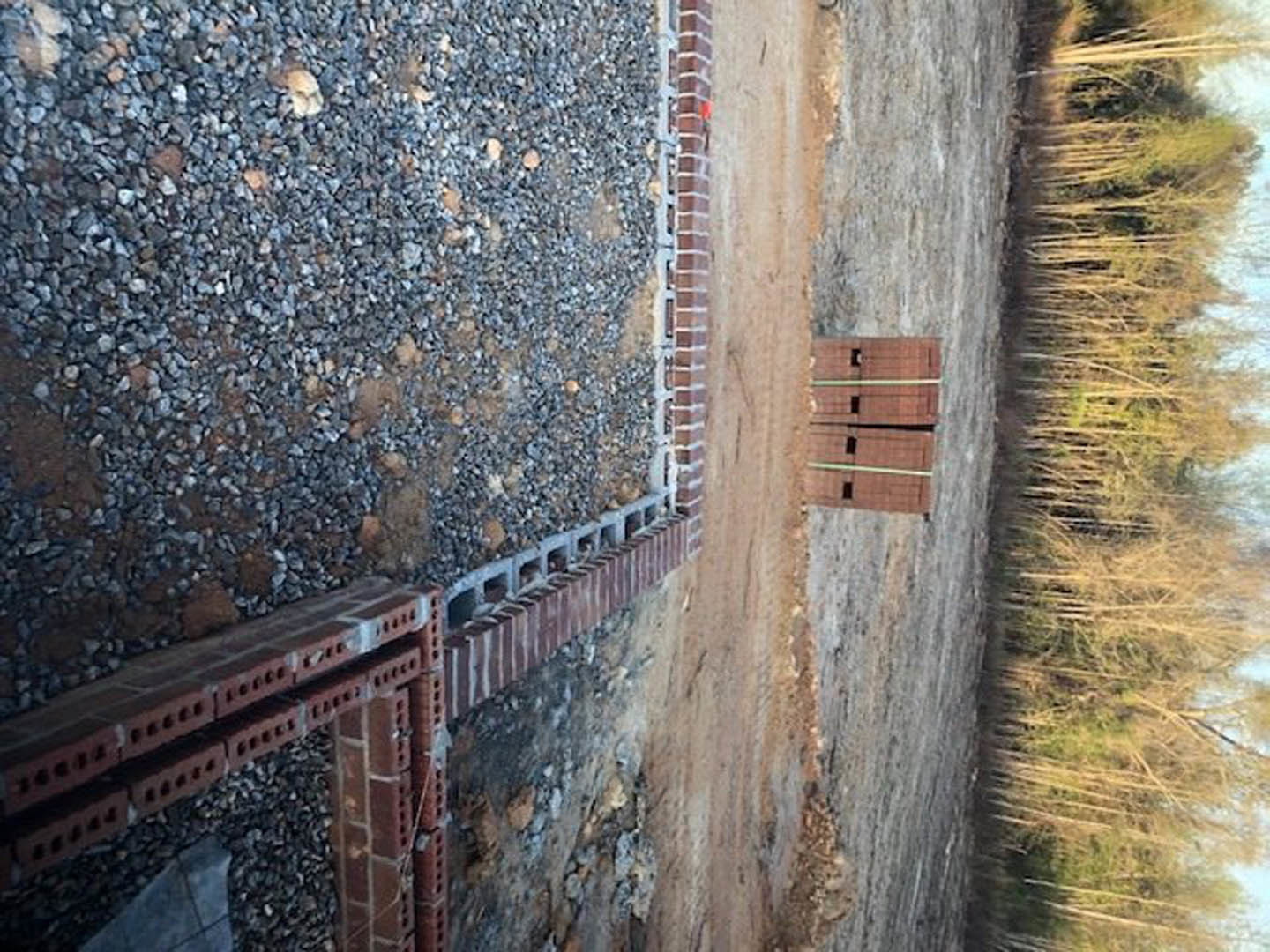 Red brick wall with small holes bordered by gravel, adjacent to a modern building, surrounded by dirt and scattered trees