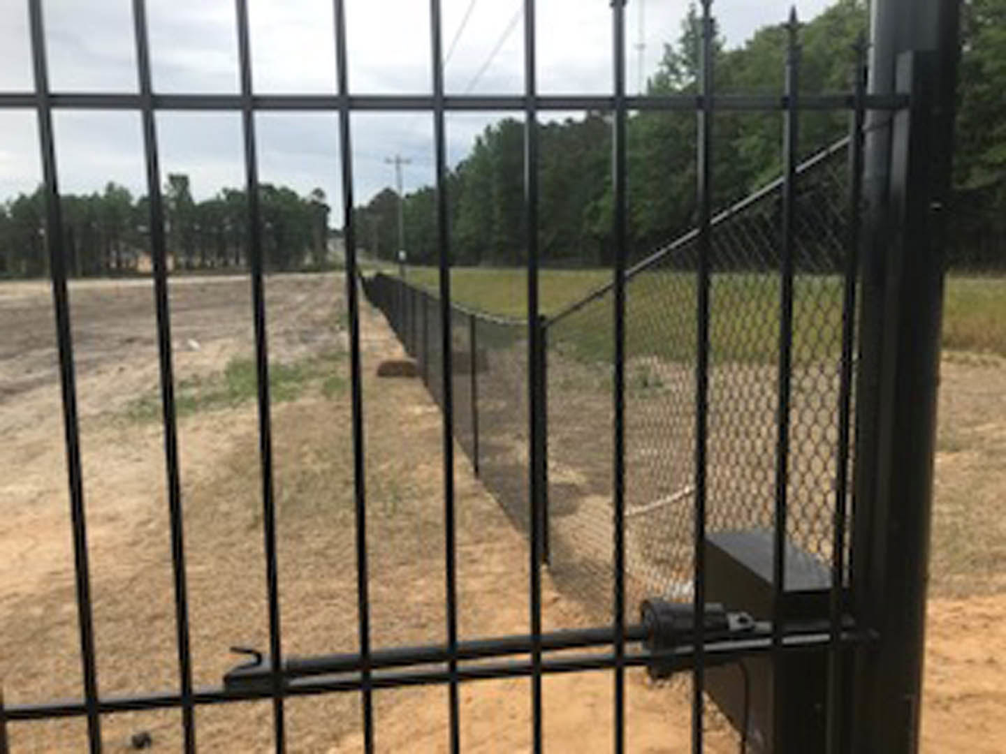 Black iron mesh fence with chain link panels enclosing grassy field, trees and blue sky in background, exterior of residential building visible.