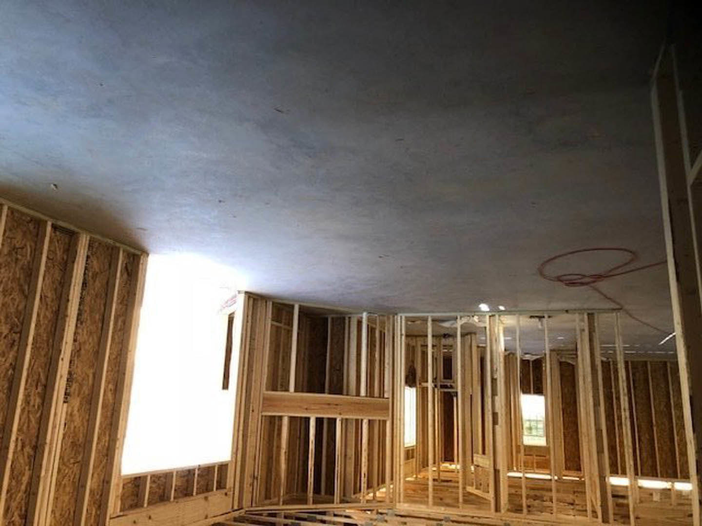 Spacious room featuring large windows, white plaster ceiling with red construction lines, exposed wooden beams, and a door frame against smooth white walls.