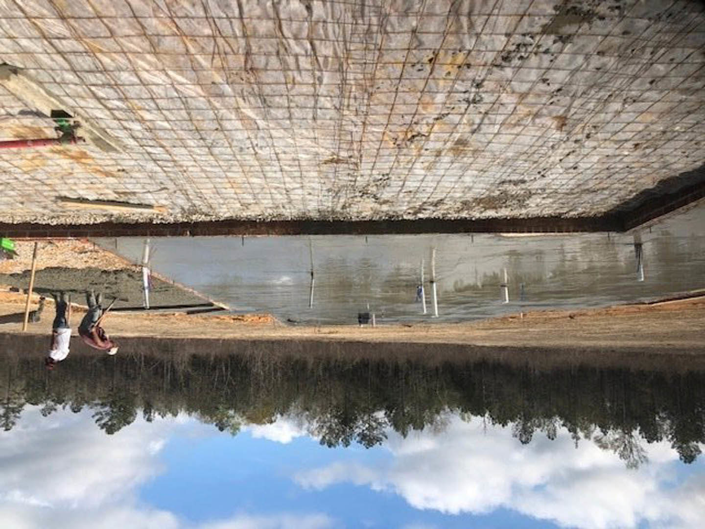 Modern lakeside home with expansive windows, wood siding, and a man standing on a dock over calm water reflecting trees and sky.