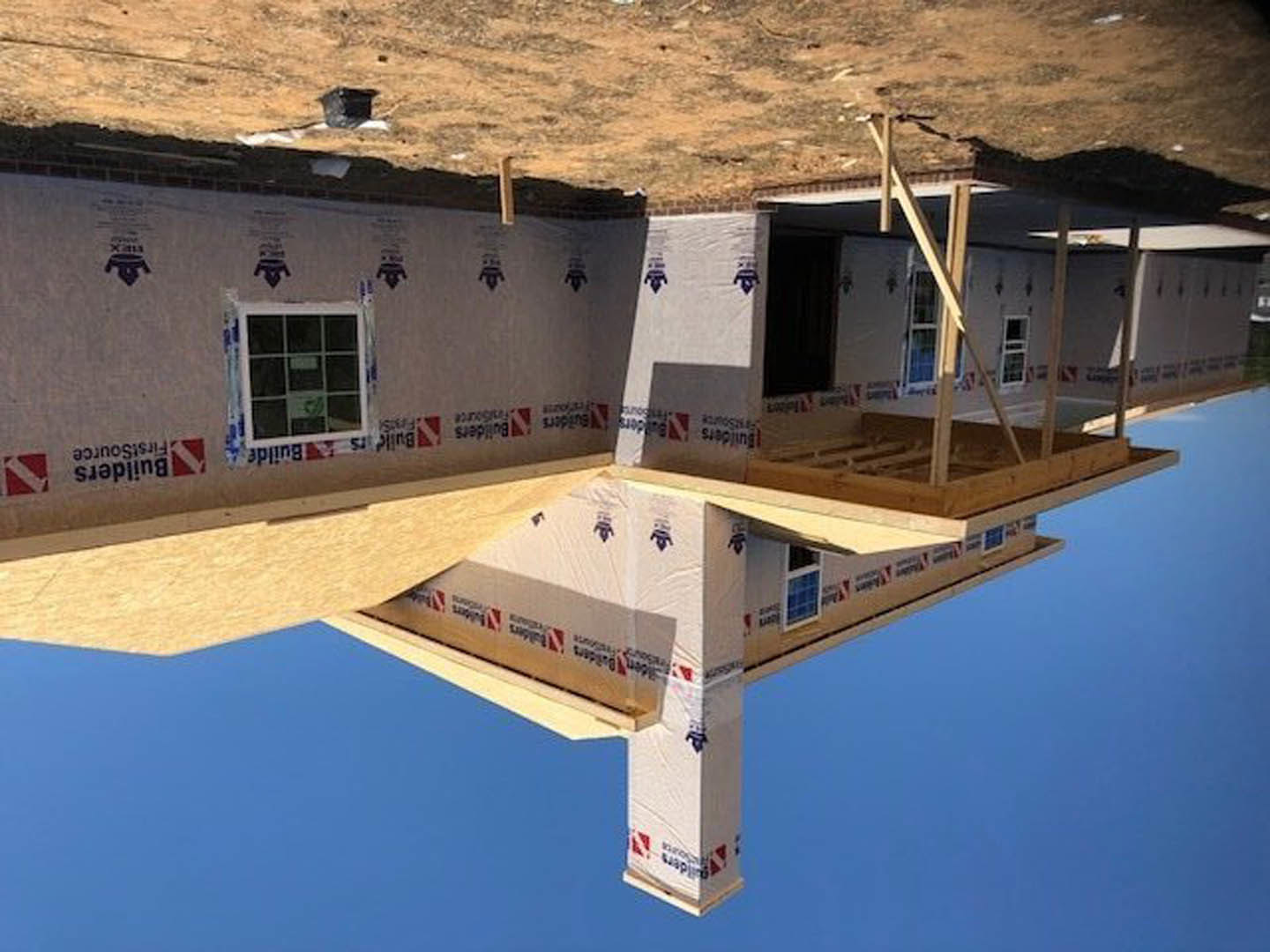 Framed custom home under construction with exposed composite materials, square window opening, dirt foreground, and clear blue sky