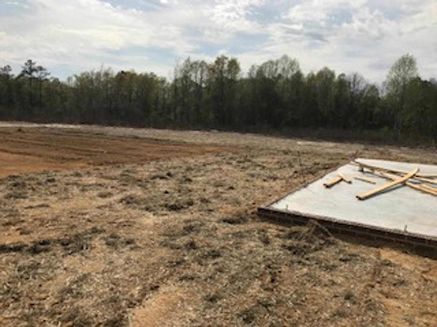 Concrete foundation slab surrounded by dirt field, scattered boards, and mature trees under cloudy sky