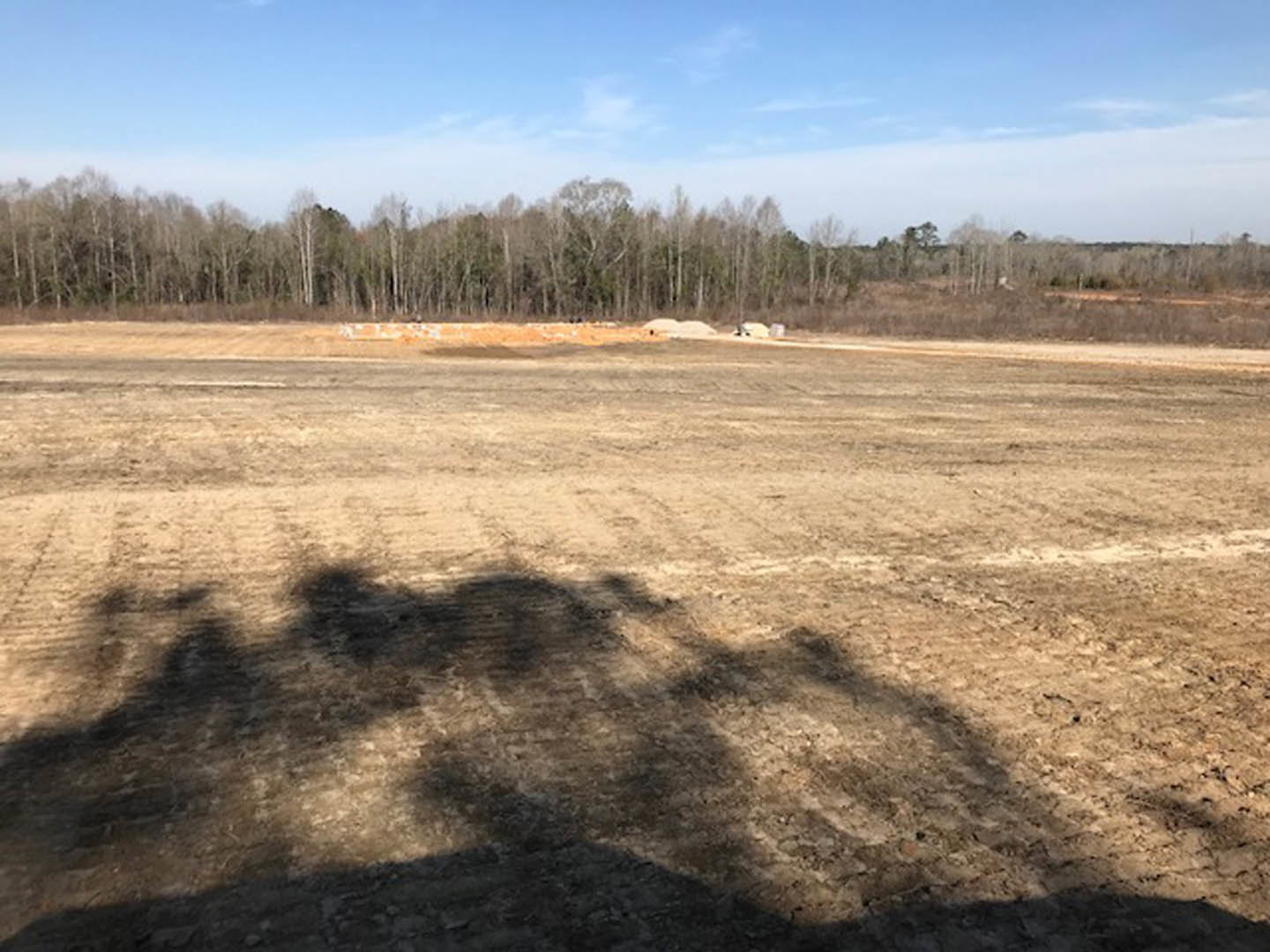 Expansive dirt field bordered by dense trees under a partly cloudy blue sky, tree shadows cast across the ground