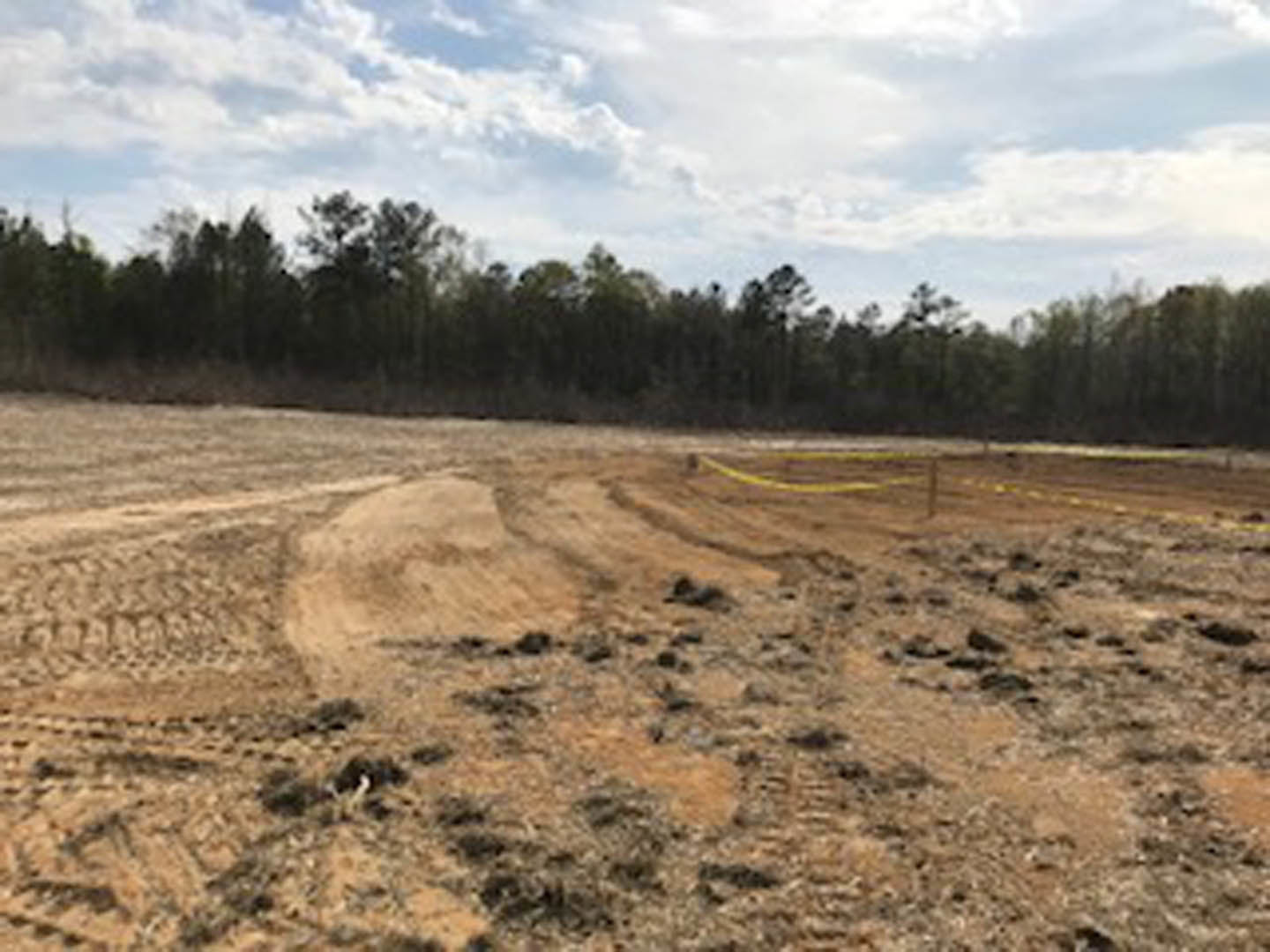 Dirt field bordered by a yellow line, distant cluster of trees, blue sky with scattered clouds, partial view of a hill, blurry fence in foreground