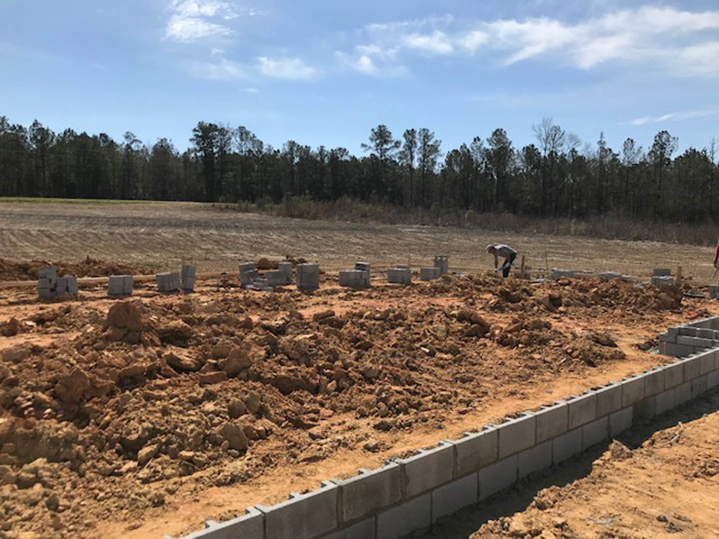 Man bending over near stone foundation at custom home construction site, surrounded by soil, trees, and cloudy blue sky