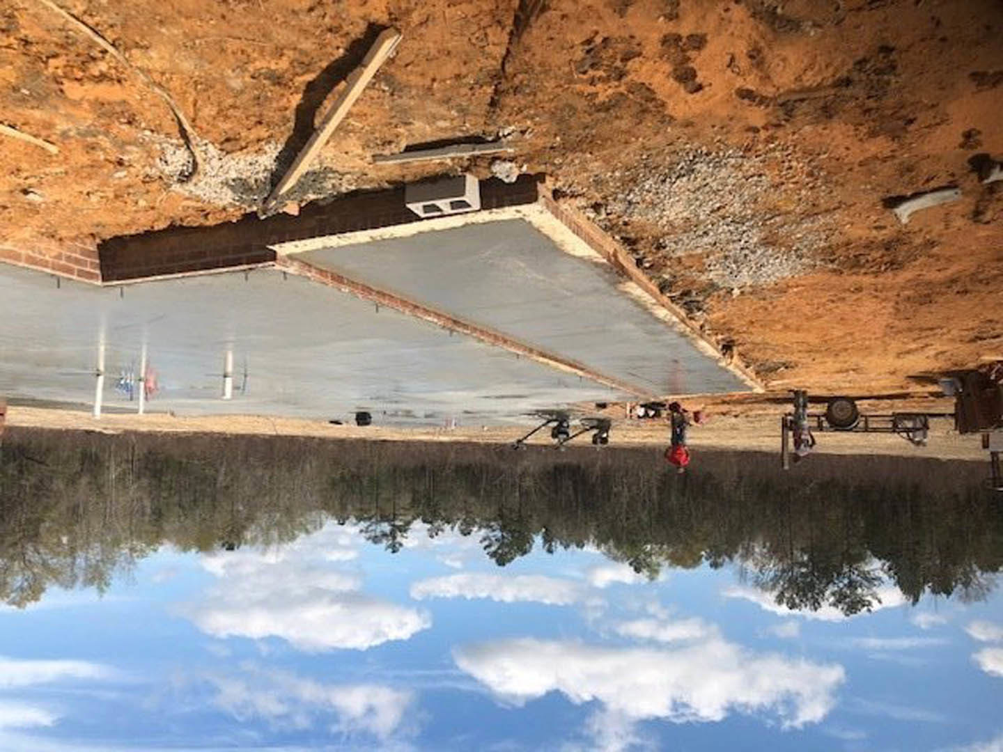 Concrete slab foundation surrounded by dirt, wooden beam resting on ground, partially built concrete wall with opening, workers present, cloudy sky reflected in water nearby