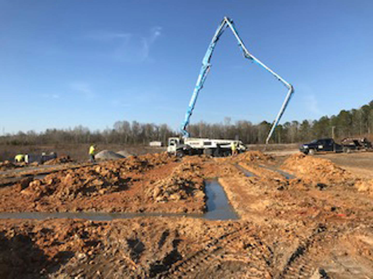 Crane lifting a white truck over dirt patch with puddle, surrounded by trees and construction equipment under open sky