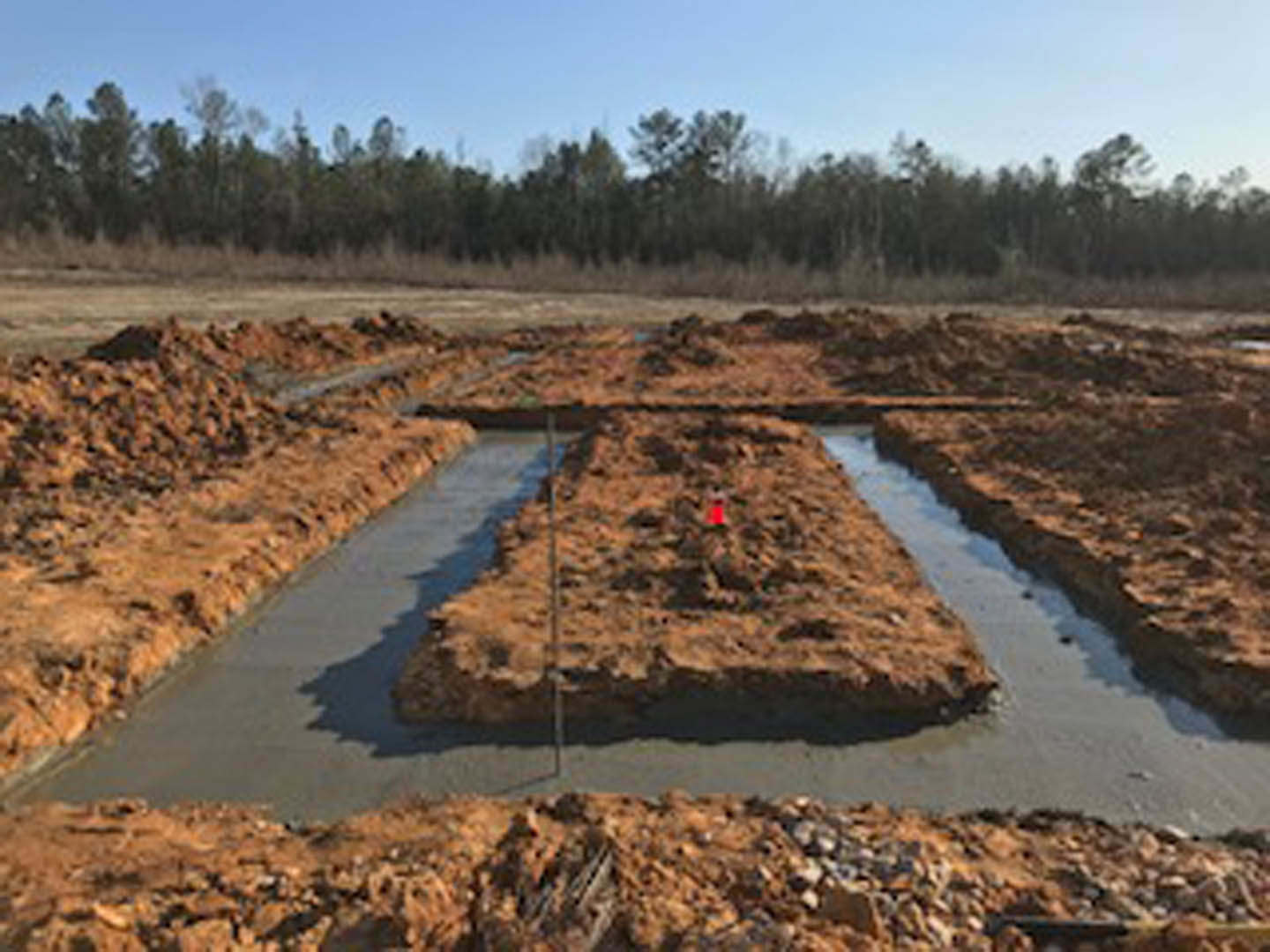 Rectangular dirt excavation bordered by trees, with a shallow stream of water running through the site and a red construction cone placed on a hill.