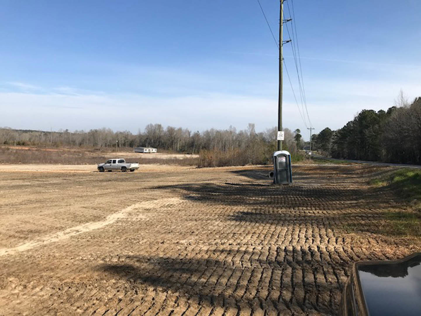 White pickup truck parked on dirt driveway beside grassy field, surrounded by tall trees under blue sky with scattered clouds