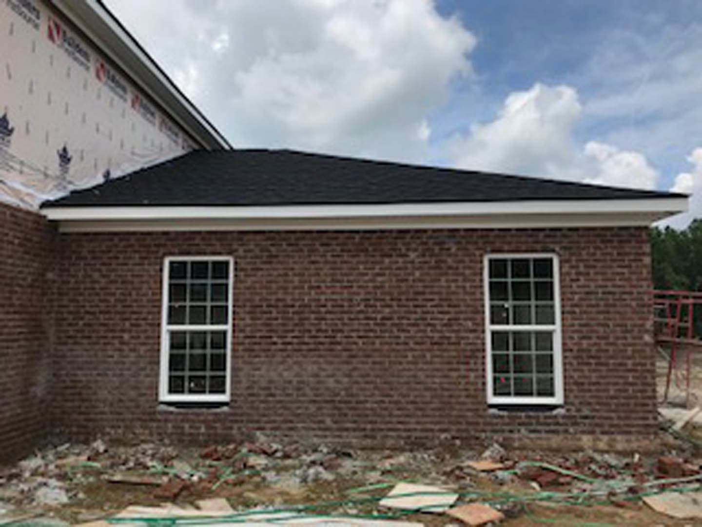 Red brick exterior wall with white-framed window, construction materials visible, blue sky and clouds overhead, person walking in foreground