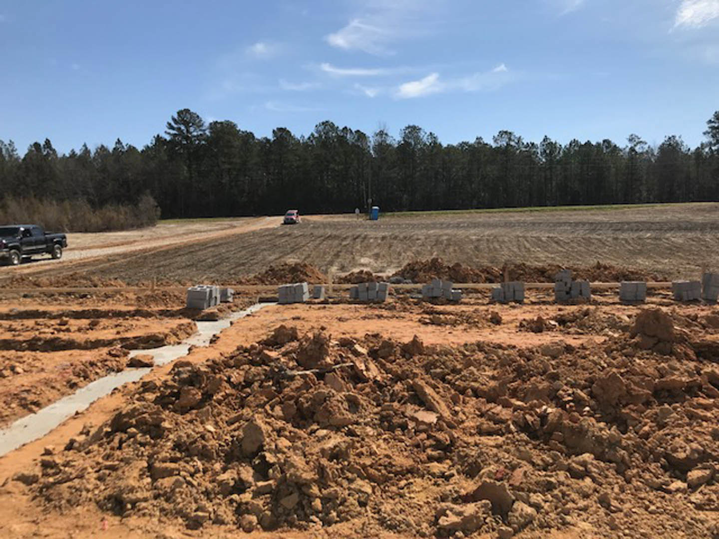 Dirt field with scattered bricks and a black truck parked on a dirt road, group of trees in the background under a blue sky with clouds
