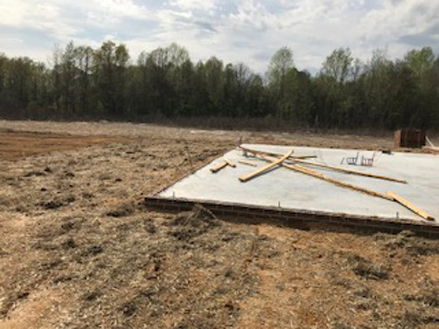 Concrete slab foundation with stacked wooden planks, surrounded by grassy land and trees under a cloudy sky