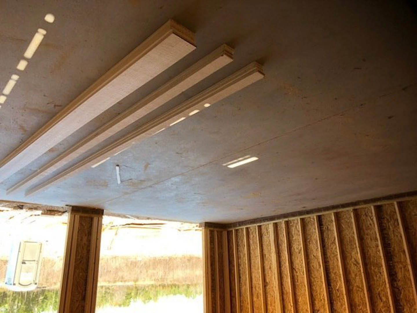 Ceiling with exposed wood beams, large window with white curtains, and light-colored walls in a corner of a room.