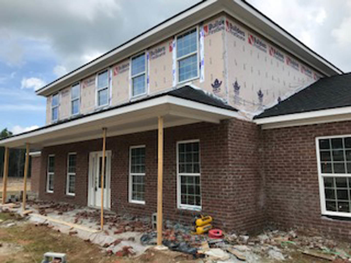 Red brick house under construction with white-framed windows, partially finished roof, and construction materials visible in the foreground