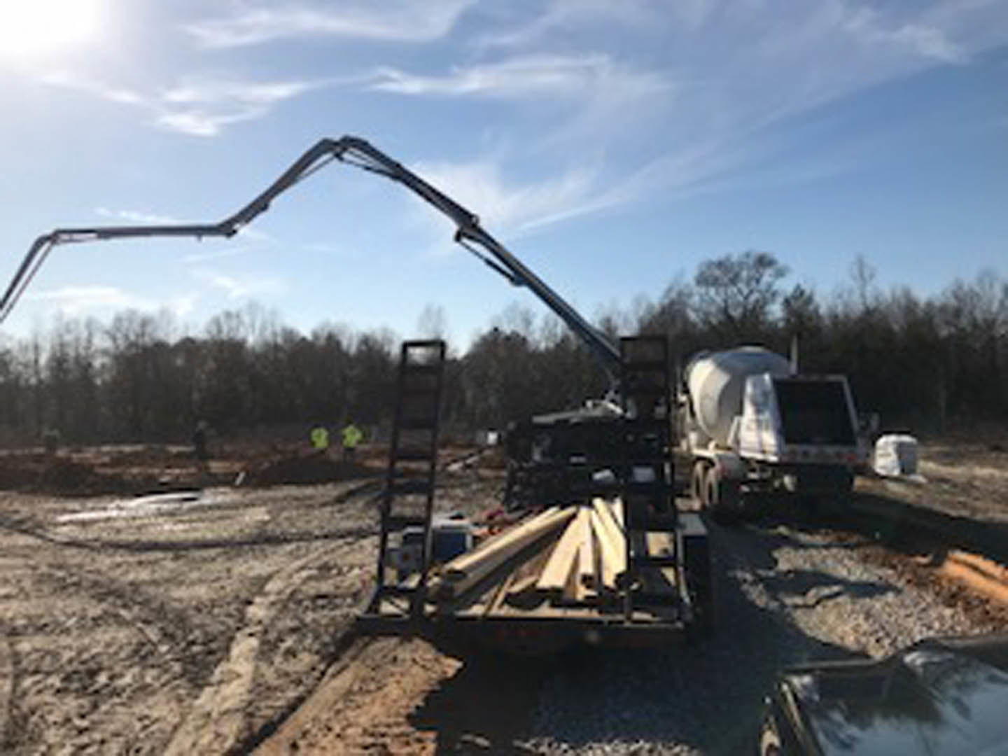 Construction site with a crane truck positioned on soil, surrounded by trees and sky, concrete mixer and other heavy equipment visible in the background
