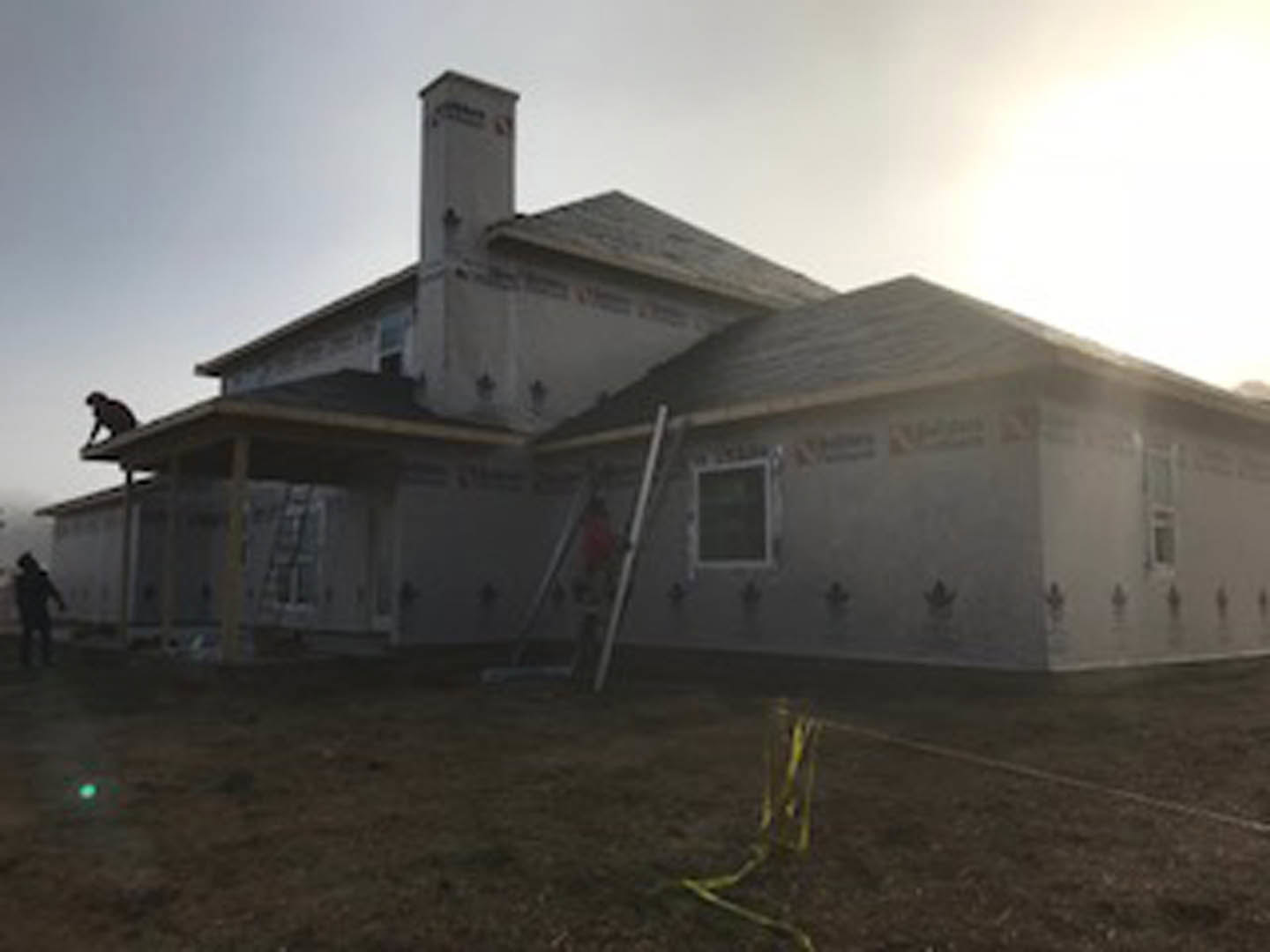 Framed house under construction with exposed wood, ladder leaning against exterior, cloudy sky overhead, grassy ground with yellow tape marking area
