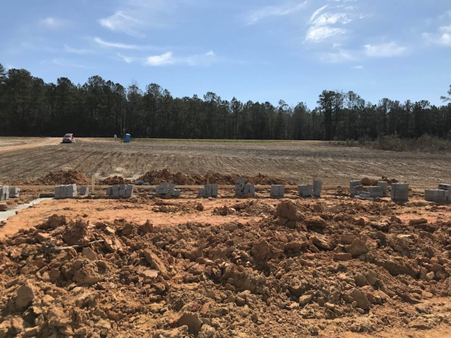 Dirt field with scattered piles of bricks and rocks, bordered by trees under a partly cloudy blue sky