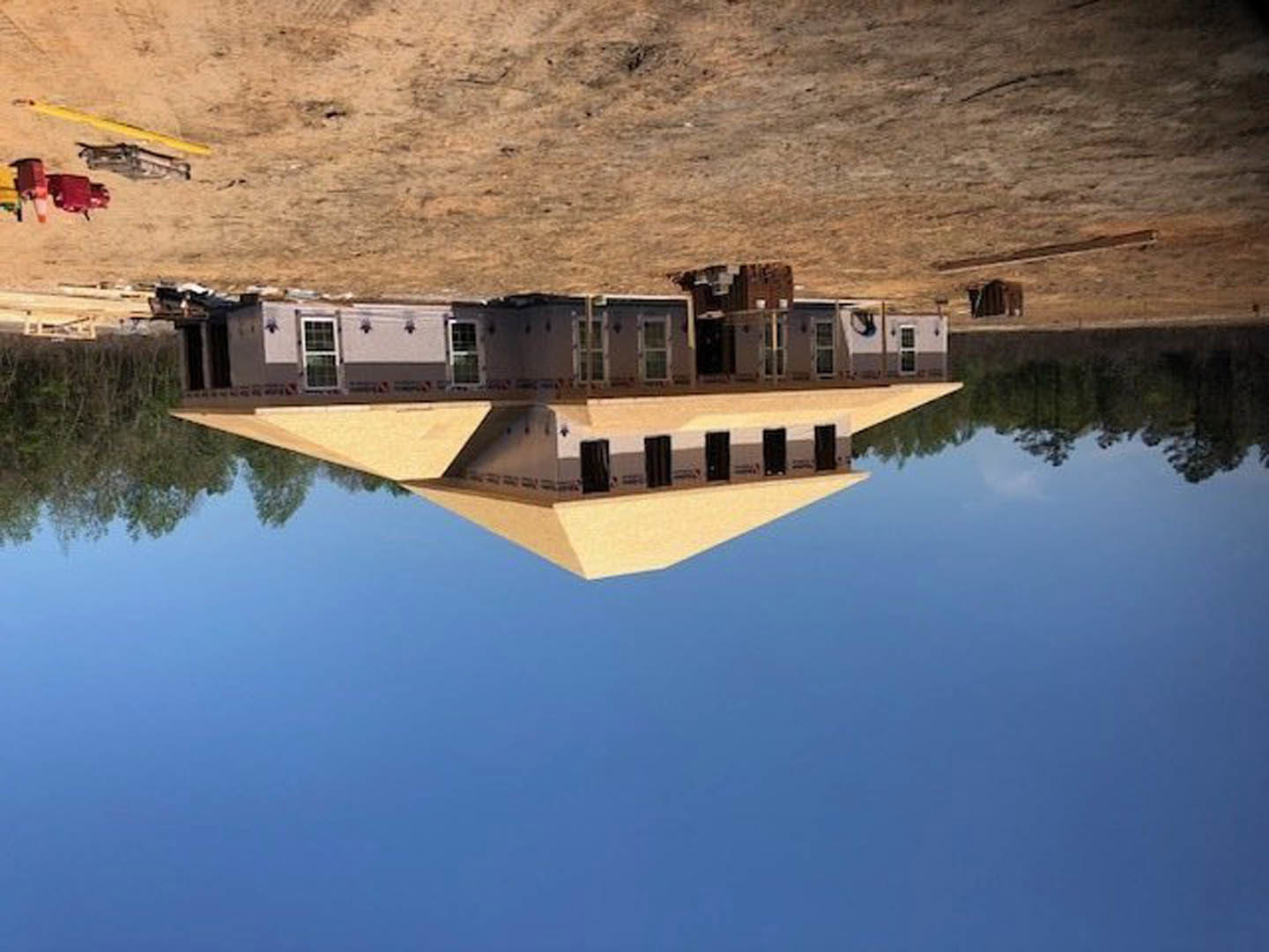 Partially built house with exposed framing and plywood sheathing, situated beside a calm lake with mountain backdrop, brown dirt construction site in foreground, bridge spanning