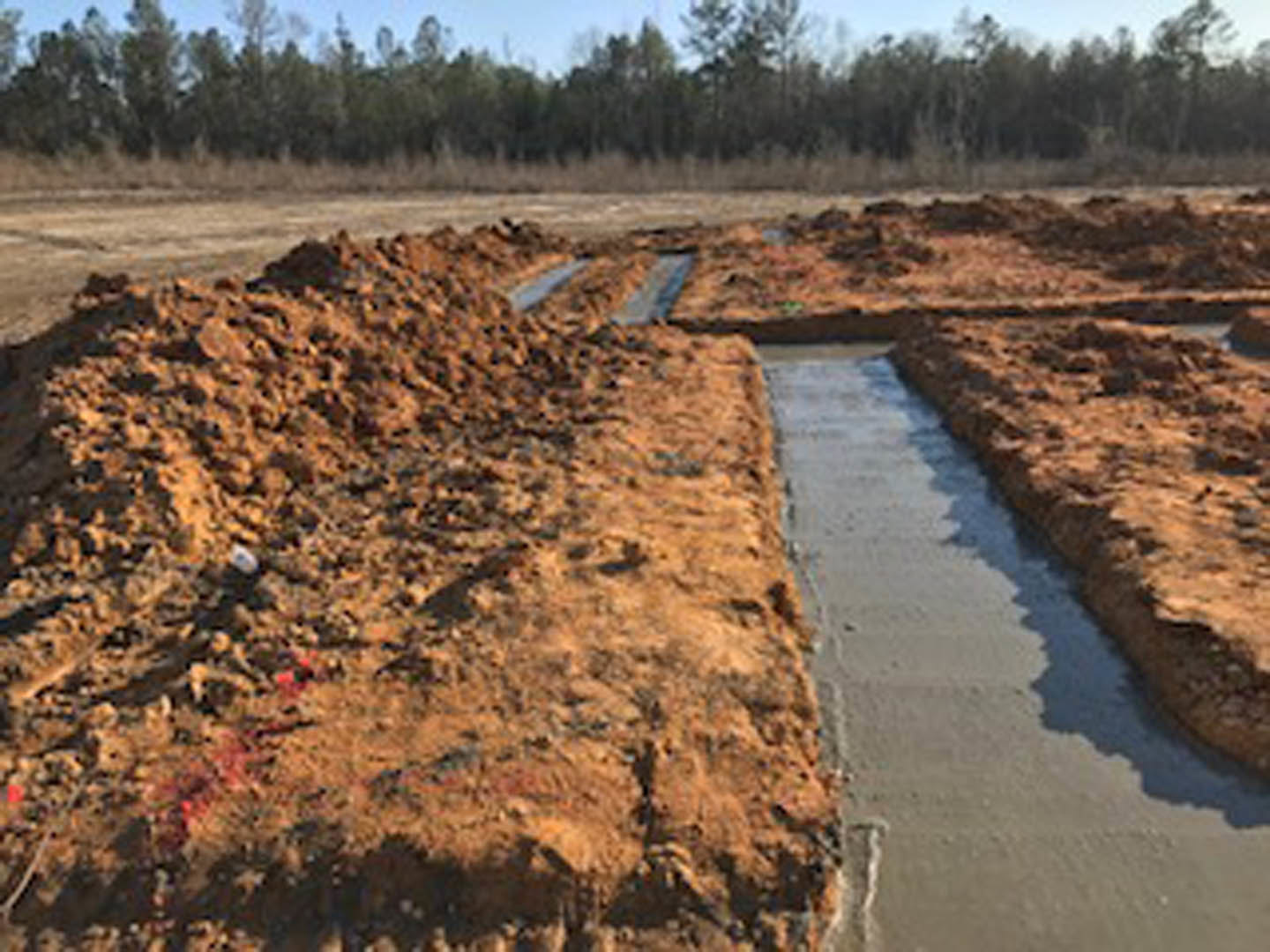 Exposed soil and dirt with several water pipes laid across a residential construction site, surrounded by trees and plants under an open sky
