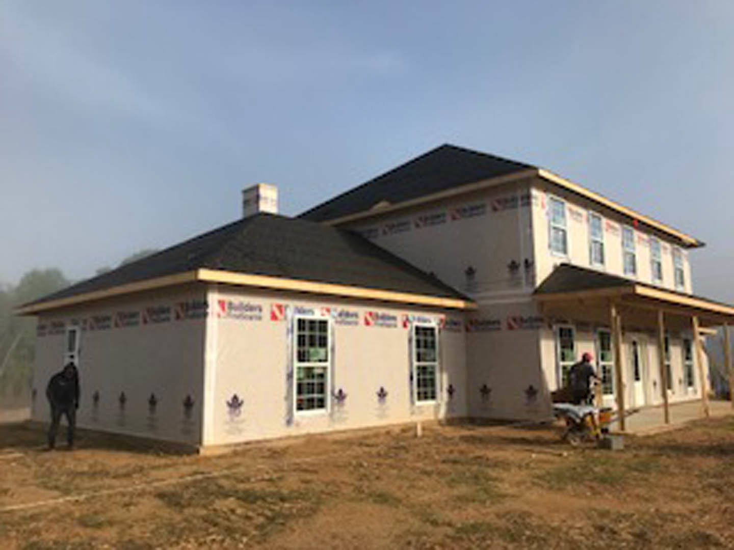 Two-story house under construction with exposed framing, black roof, white-trimmed windows, and a man standing on brown grass in front