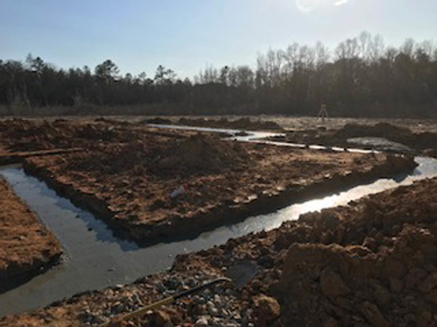 Narrow river winding through a dirt field bordered by sparse trees, under a blue sky with scattered white clouds