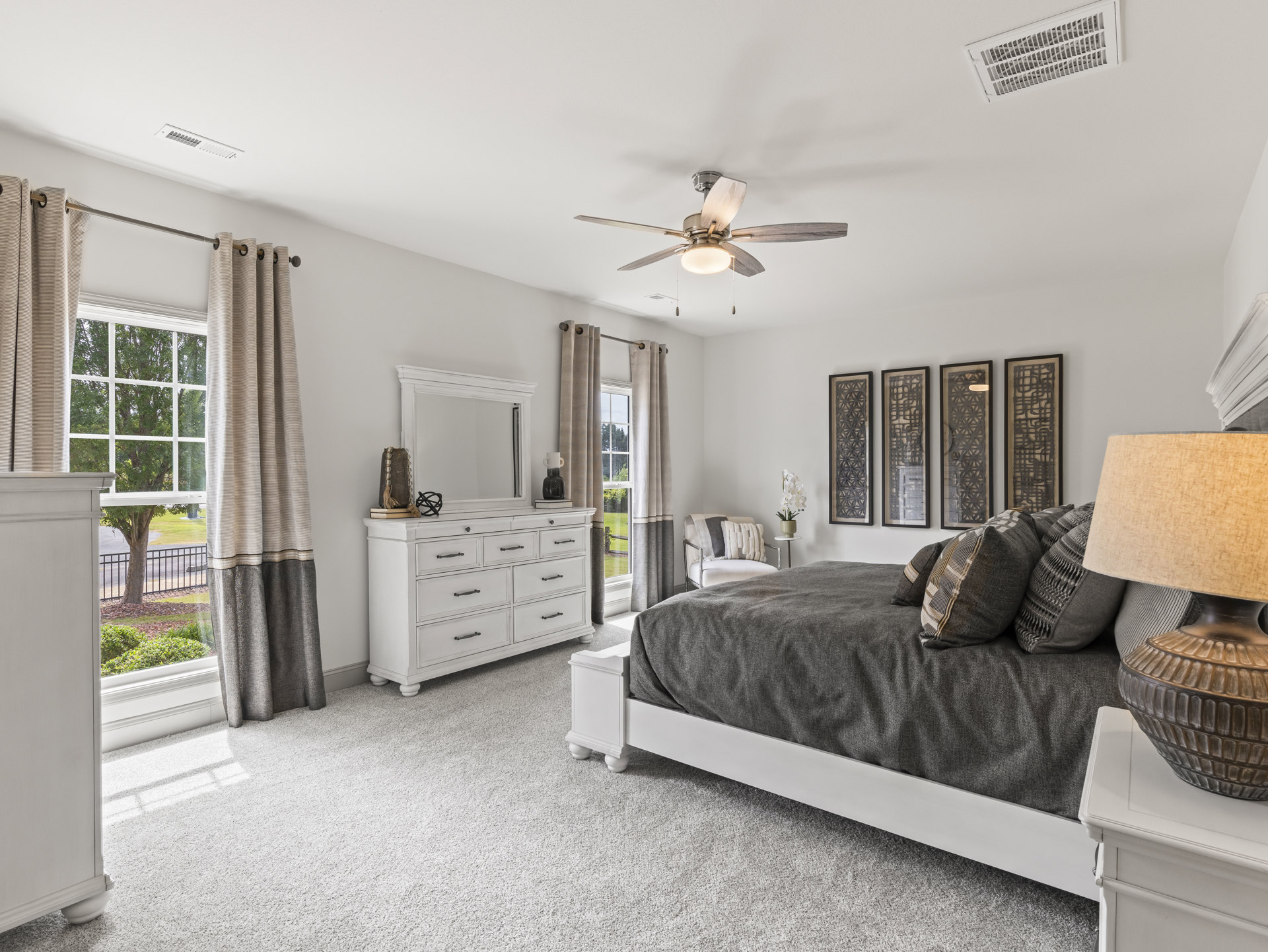 Bedroom with upholstered bed, white dresser topped with mirror and decor, ceiling fan with light fixture, framed artwork on wall, floor lamp, and light curtains.