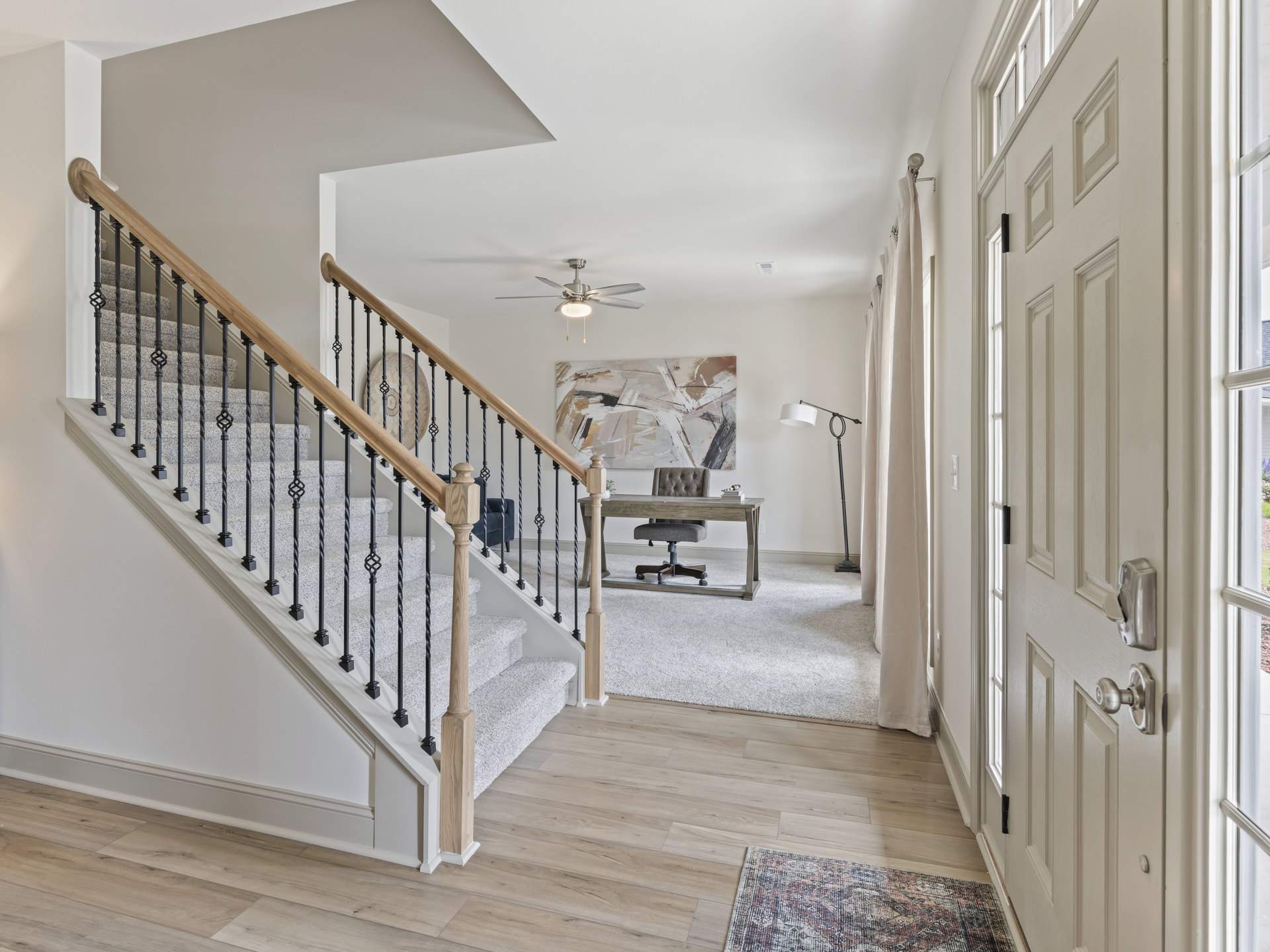 Hallway with light wood flooring, black metal staircase railing, white door, circular wall art, patterned rug, and ceiling fan with light fixture
