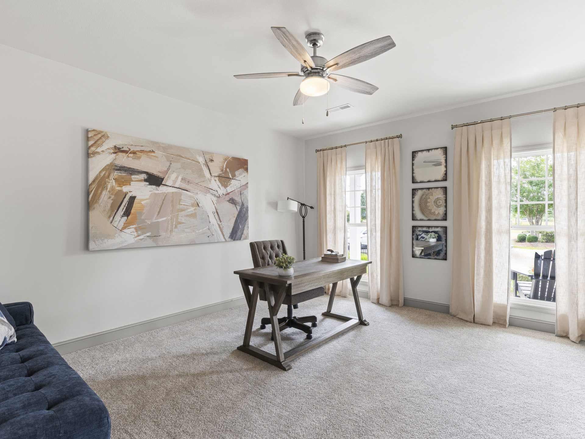 Bright home office with wood desk, white potted plant, ceiling fan with light, gray couch, and abstract painting on pale wall