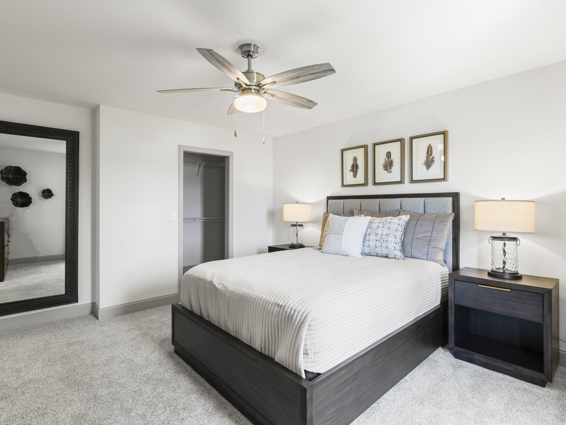Queen bed with white linens and pillows, wood nightstand with glass lamp, ceiling fan above, framed feather artwork on white wall