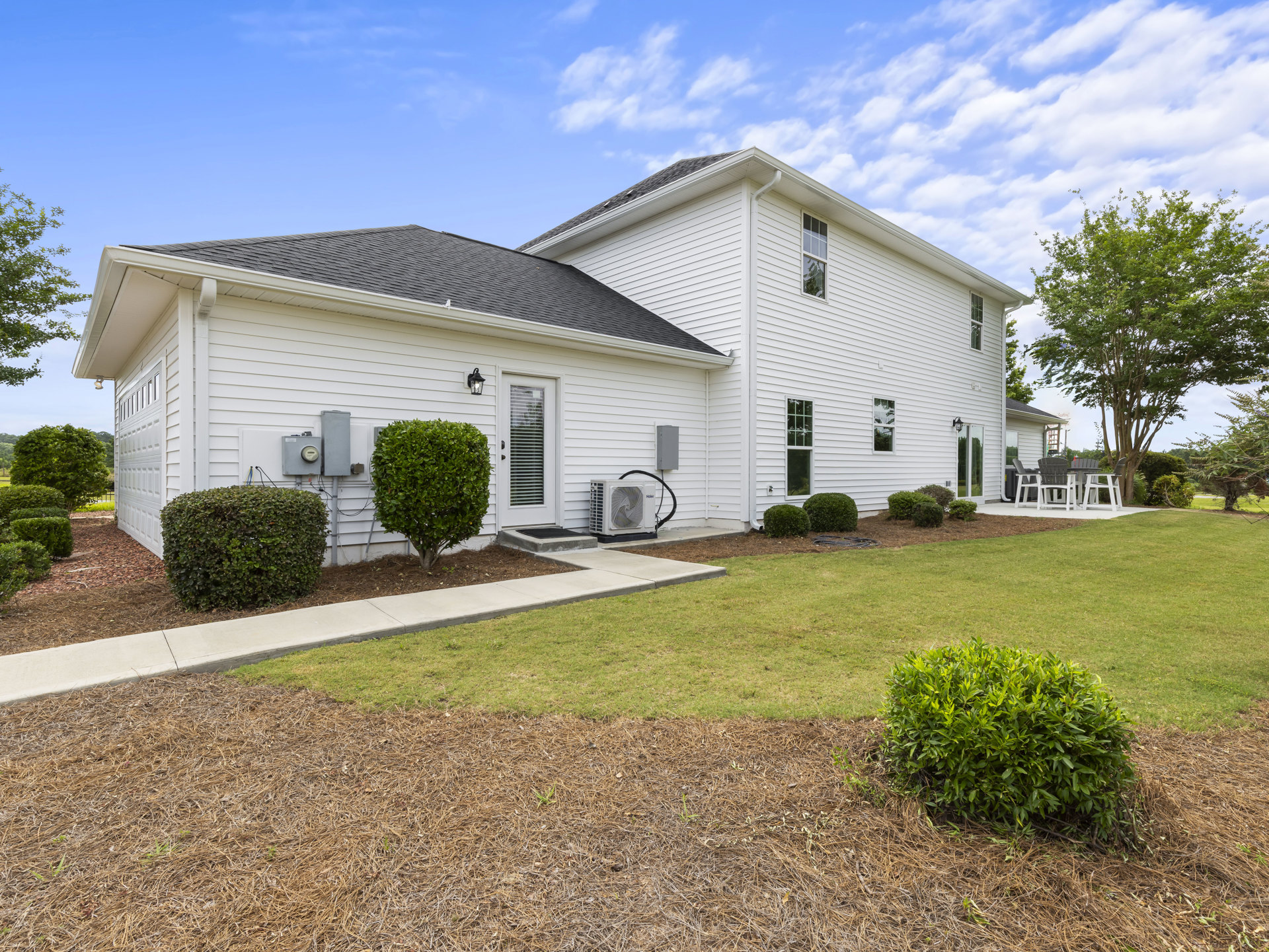 White siding house with green lawn, leafy bushes, and trees; white front door and porch with chairs; cloudy sky overhead.