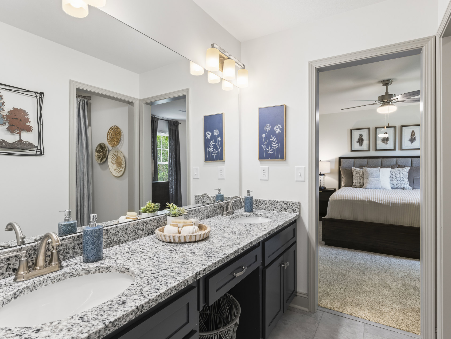 Bathroom with double sink vanity, light wood cabinetry, white countertops, bed visible in adjoining space, potted plant, decorative metal tree wall art, soap dispenser, towels and