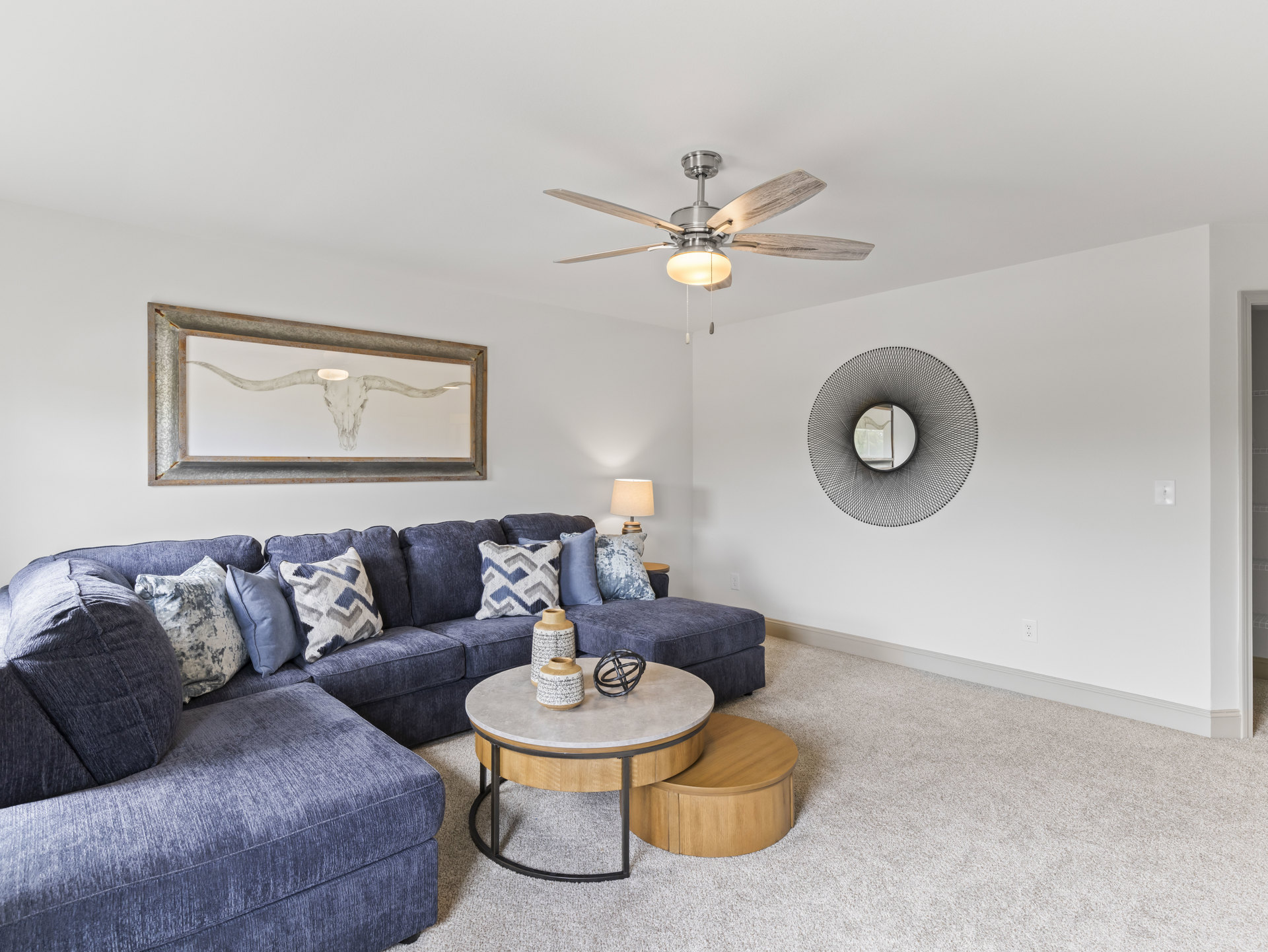 Living room with light gray couch, ceiling fan with integrated light, bull skull painting, circular wall mirror, round marble-topped table, black metal sculpture, neutral walls