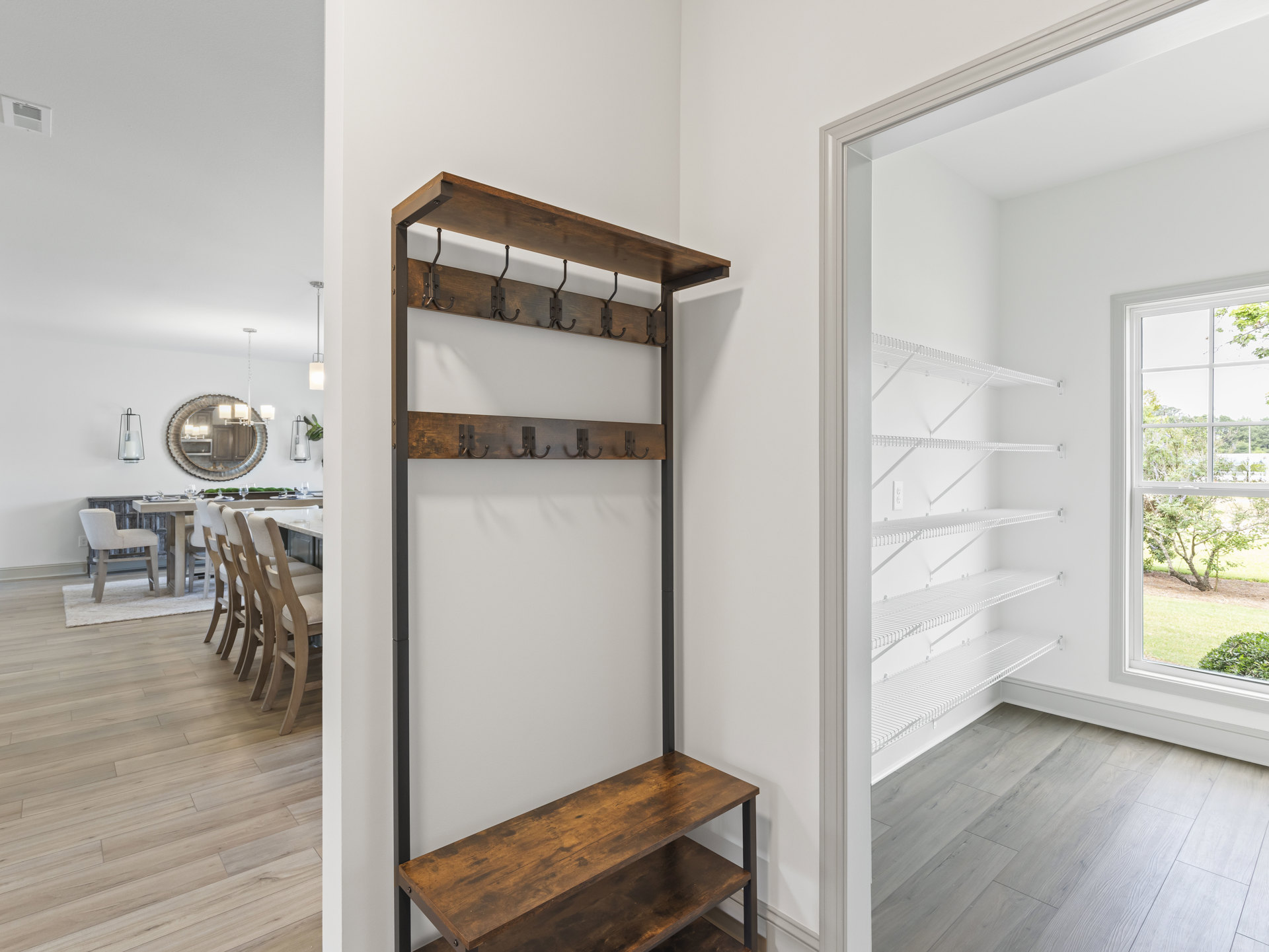 Hallway with hardwood flooring, white walls, wooden coat rack with hooks, mirror and light fixture above a shelf, chair near window overlooking trees.