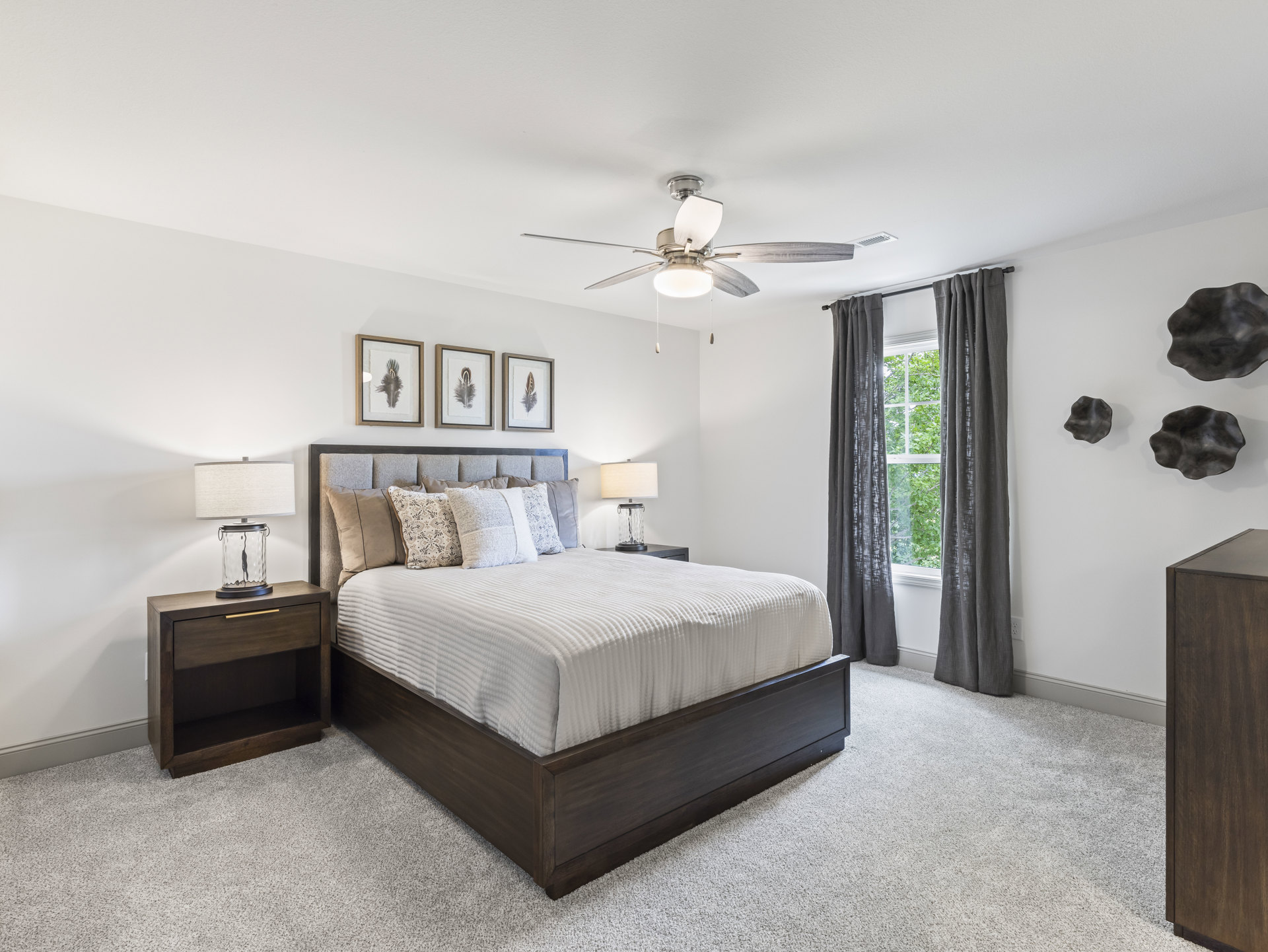 Bedroom with a ceiling fan, bed with pillows, wooden nightstand holding a glass jar, window with curtains overlooking trees, and framed feather artwork on the wall