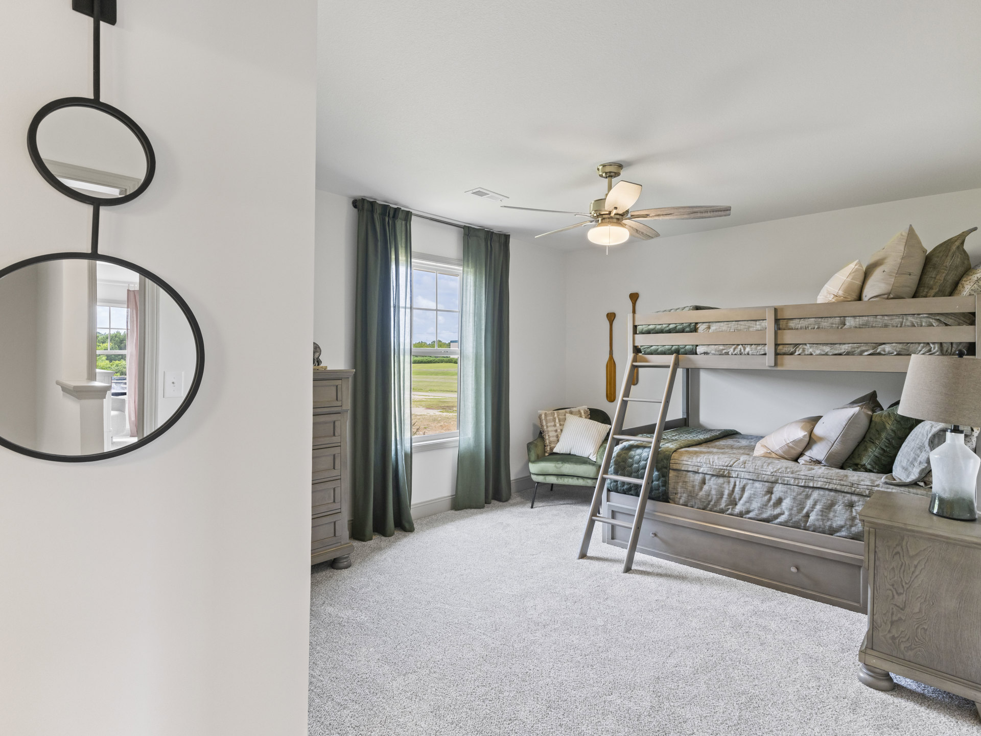 Bunk bed with white bedding and pillow, ceiling fan overhead, window overlooking grassy field, mirror mounted on wall, neutral flooring and plaster walls