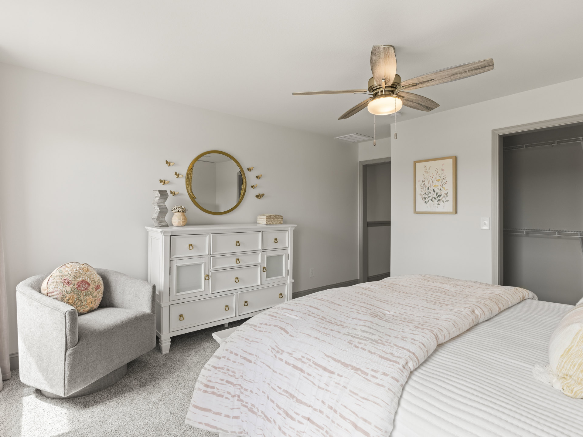 Bedroom with white dresser, white bed and bedding, round pillow on upholstered chair, floral painting in frame, wall mirror, nightstand, and ceiling fan
