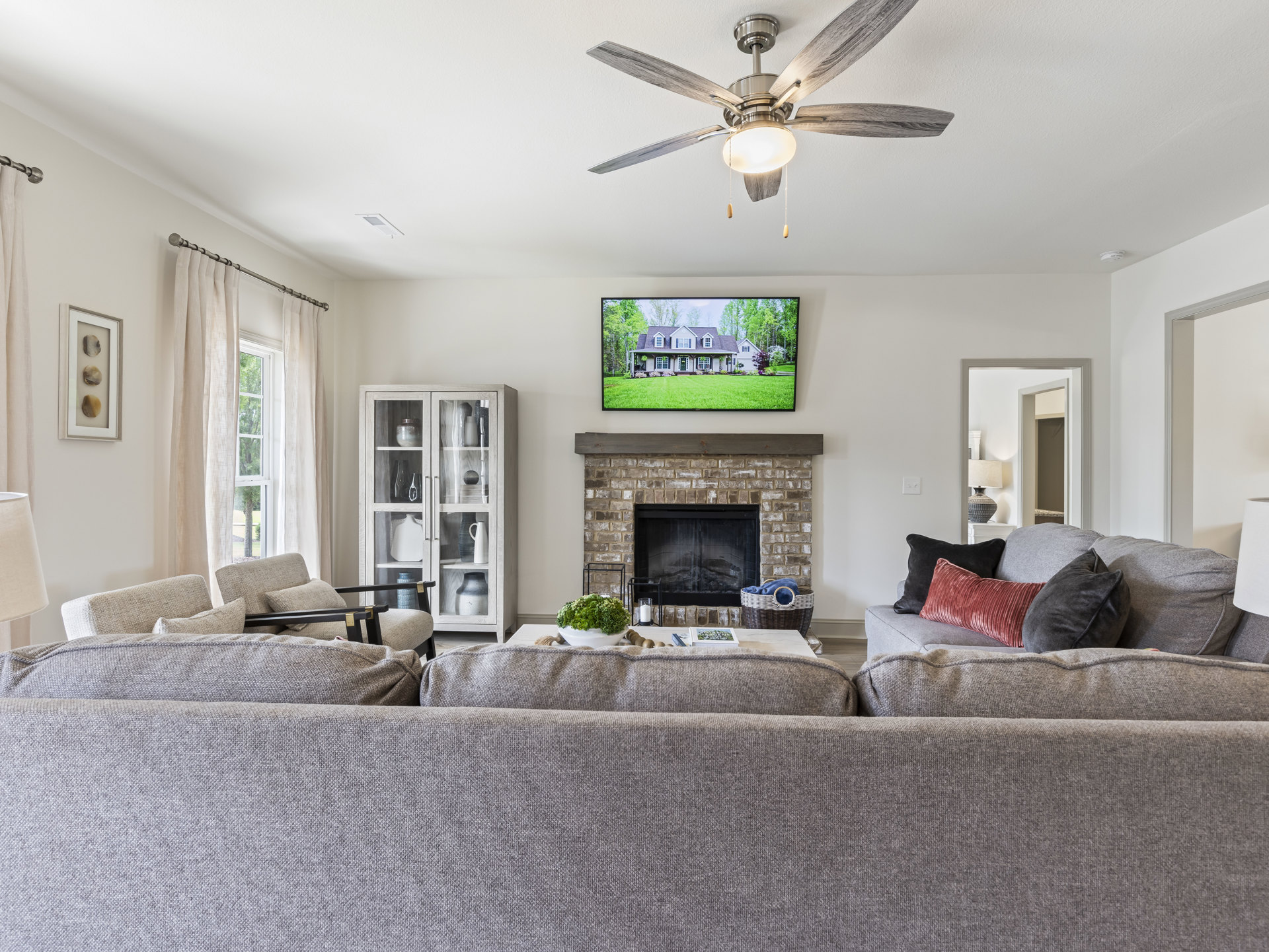 Living room with stone fireplace, gray couches, ceiling fan with light, glass-front cabinet displaying white vase, framed rock art on wall, neutral finishes throughout