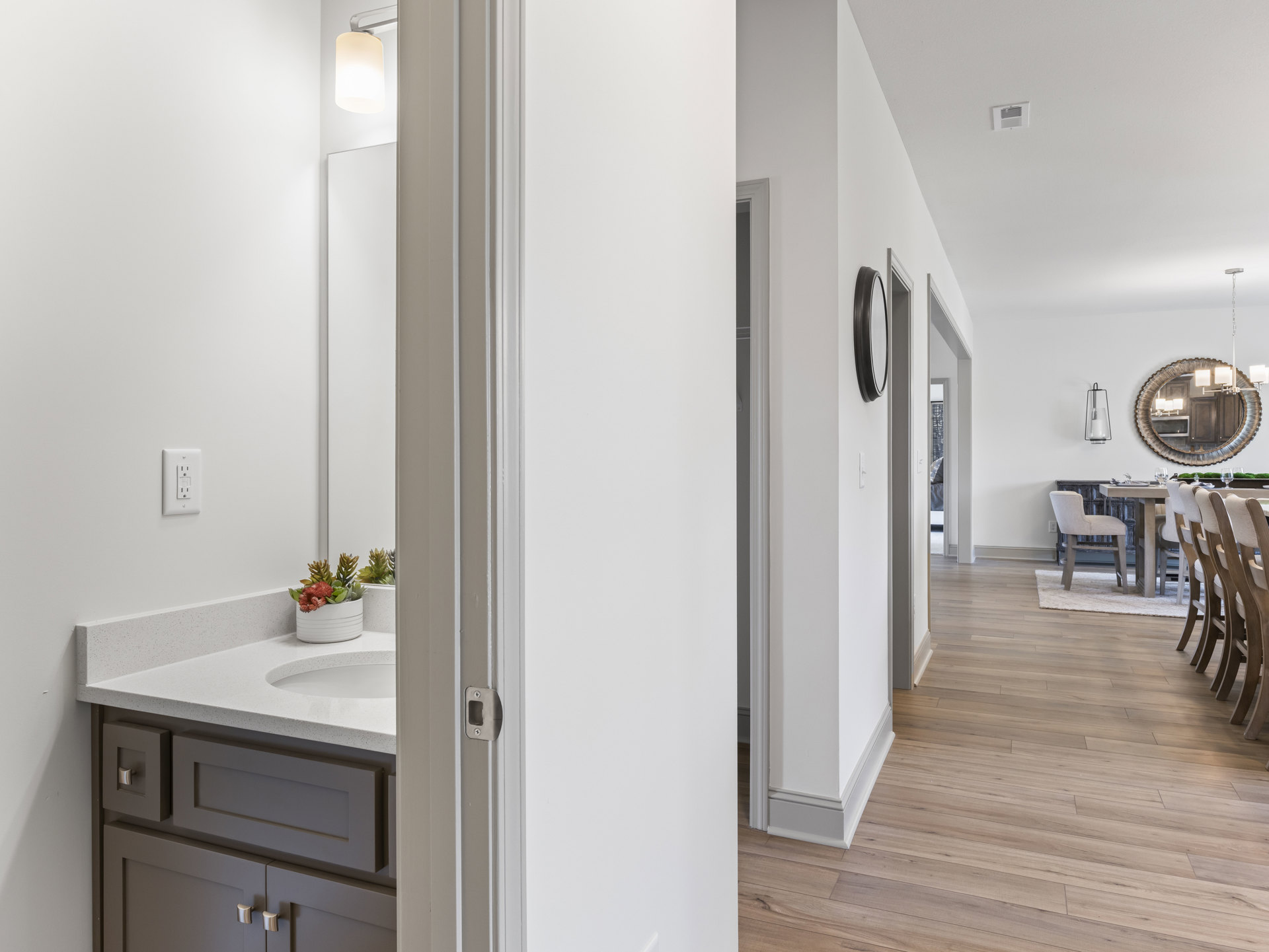 Bathroom with wood flooring, white wall outlet, mirror above sink with light fixture, potted plant on countertop, chair in corner, white cabinetry and tile accents