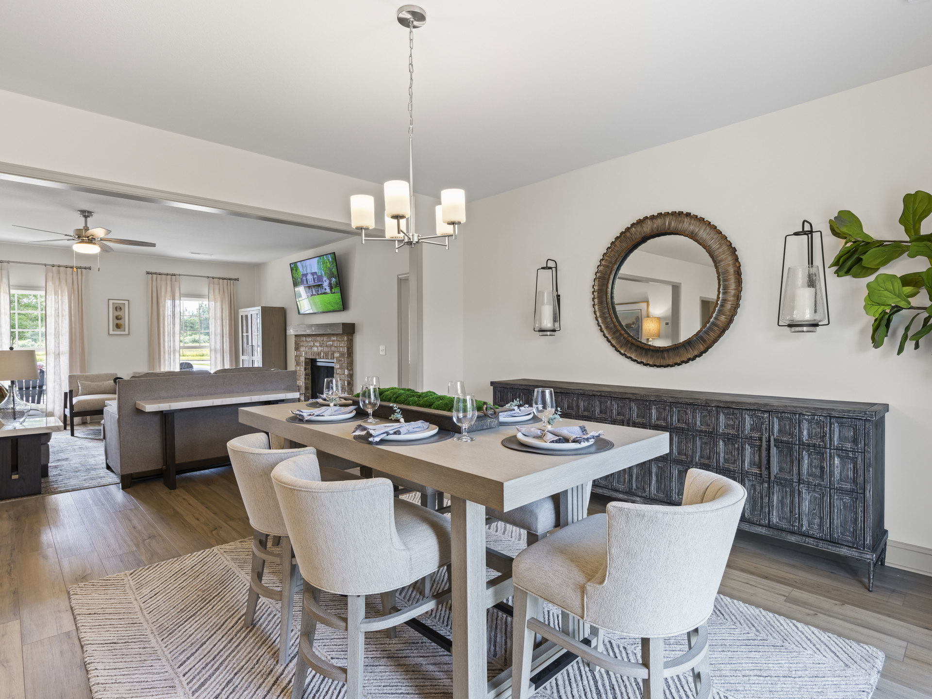 Wood dining table set with upholstered chairs, plates and glasses arranged on top, mirror and framed house artwork mounted on light-colored wall, hardwood floor and ceiling