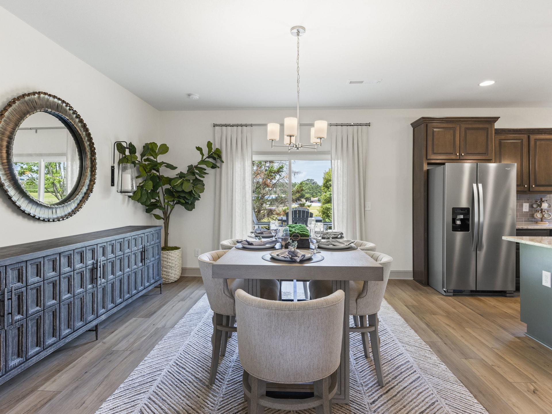 Wood dining table set with plates and glasses, surrounded by upholstered chairs, silver refrigerator with black handle in kitchen area, circular framed mirror on wall, potted plant
