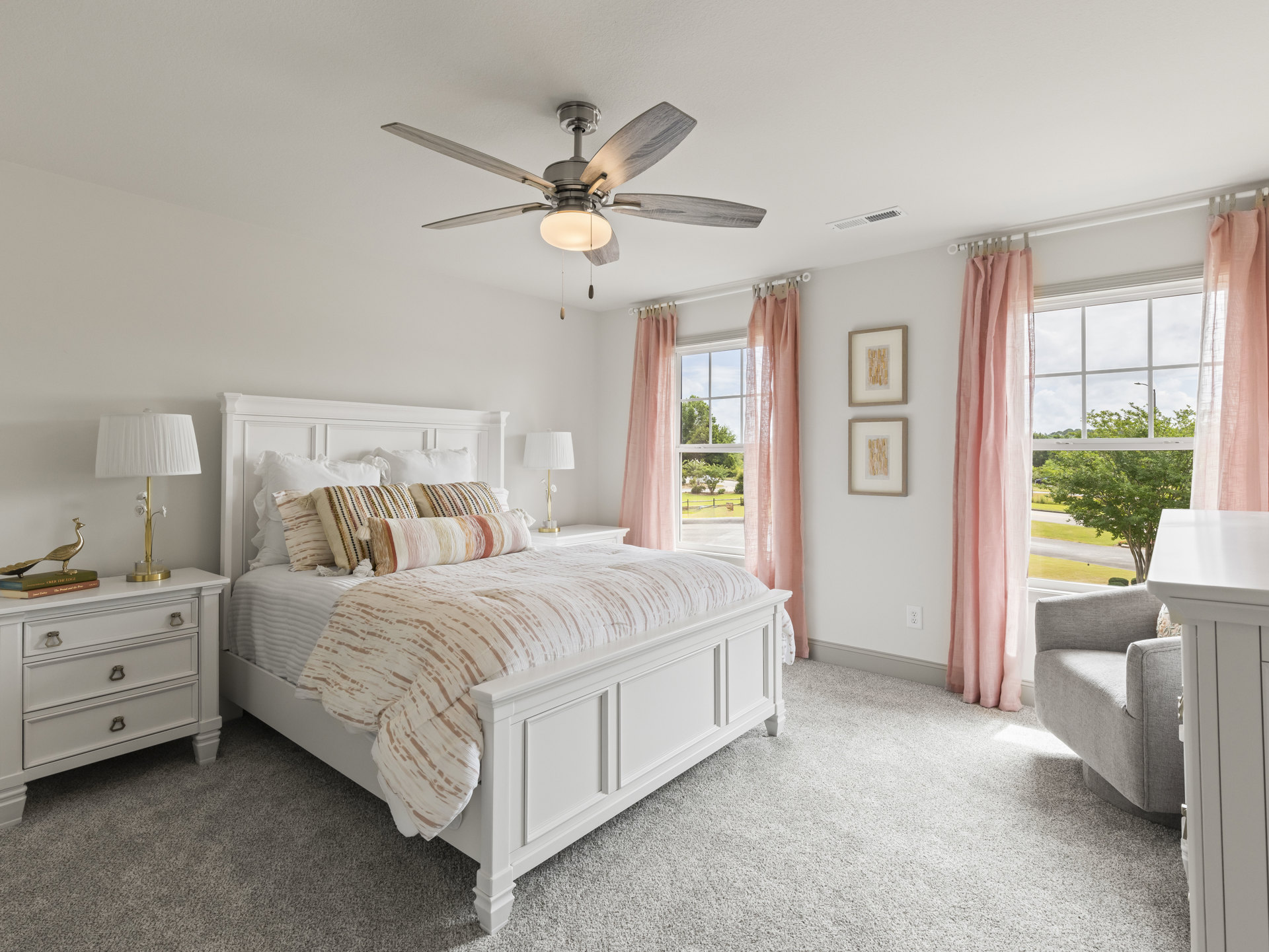 Bedroom with a ceiling fan, bed with white linens and pillows, bedside lamp with flowers, white dresser topped with lamp and books, pink curtains framing a window.