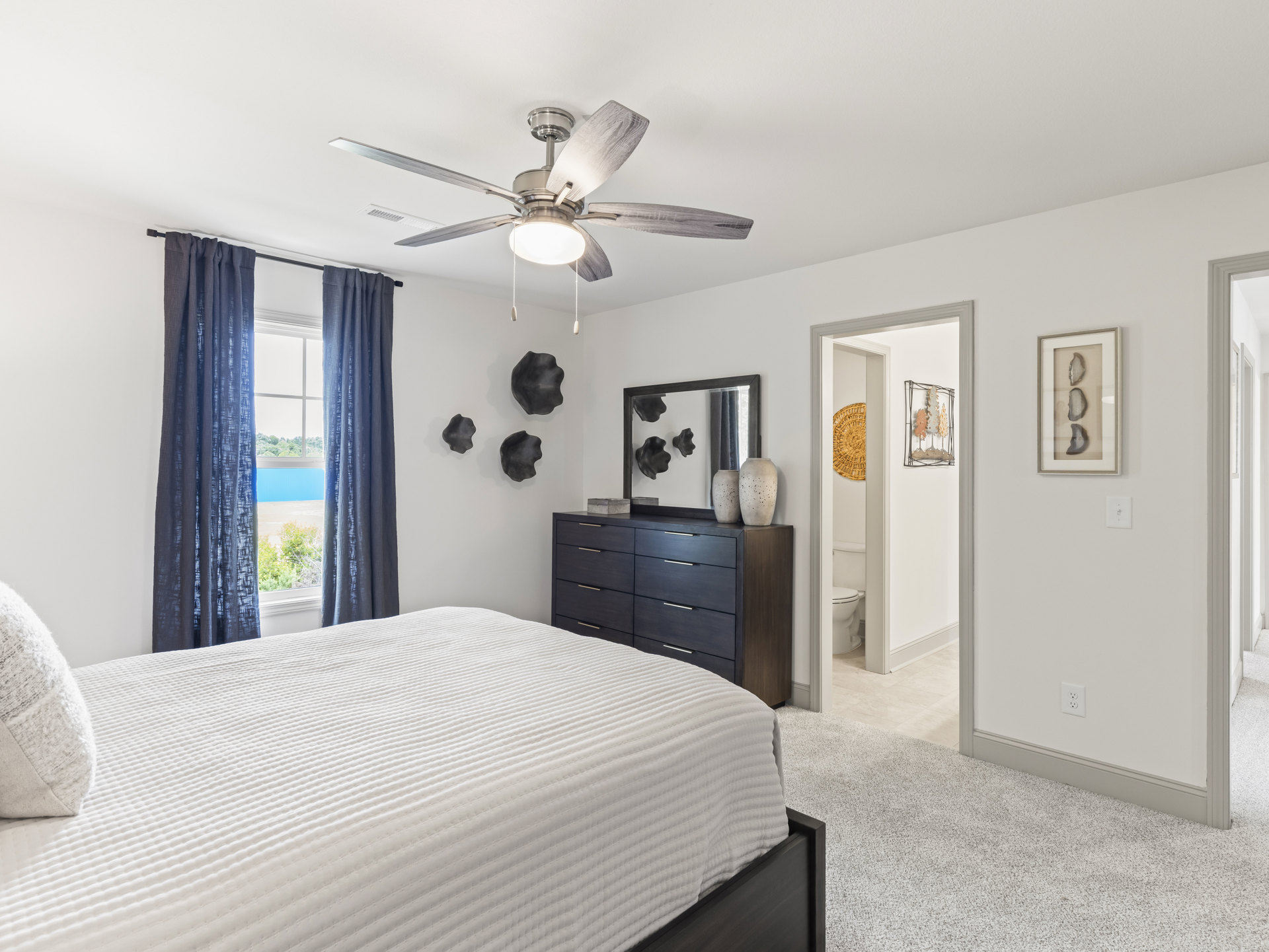 Bedroom with a ceiling fan and light fixture, bed with white blanket and pillows, framed stone artwork on wall, window with curtains showing tree outside