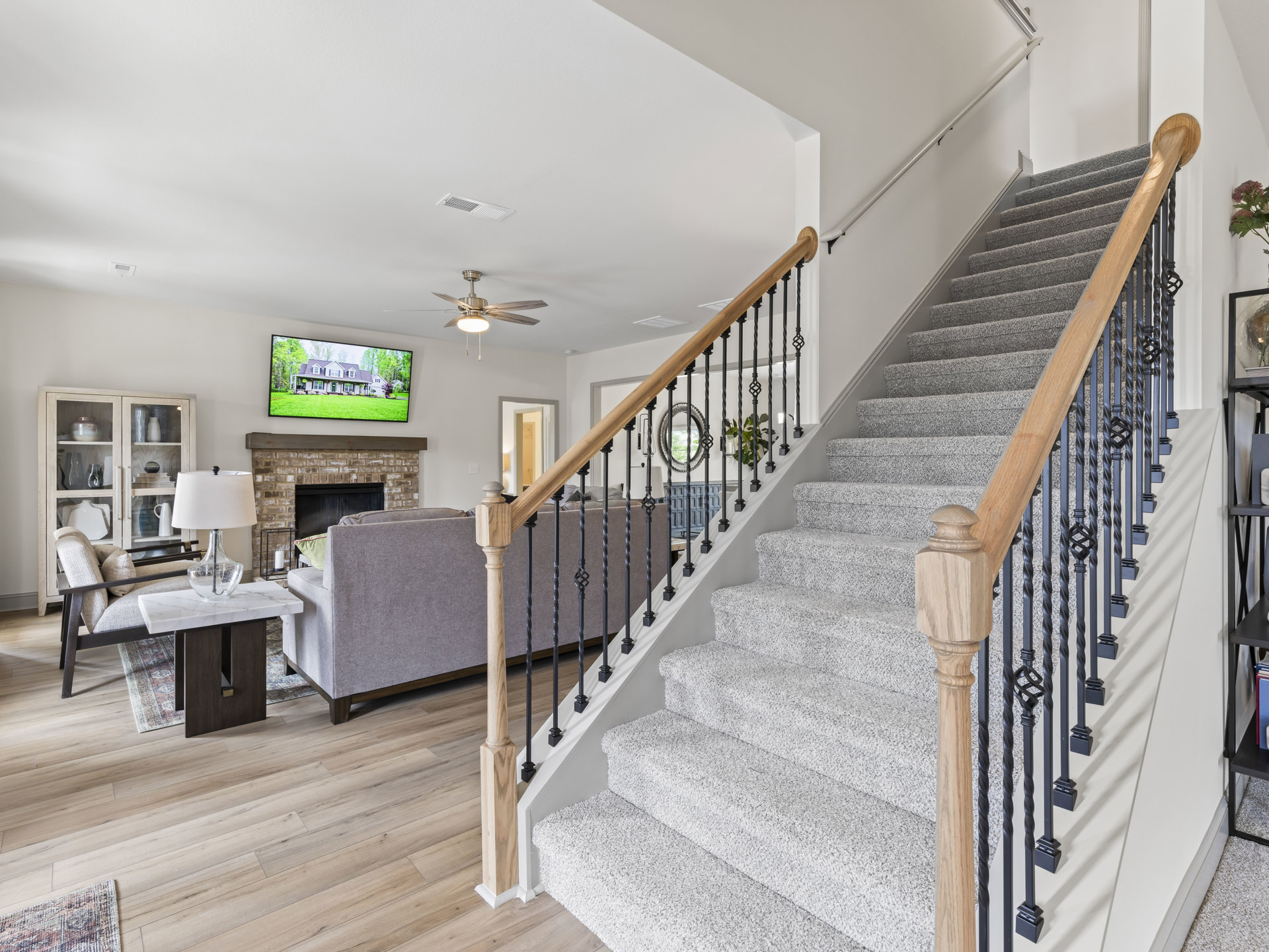 Wood staircase with wooden railing, neutral walls, mirror mounted above a couch, television screen displaying a house, light flooring, and recessed ceiling lighting in a living