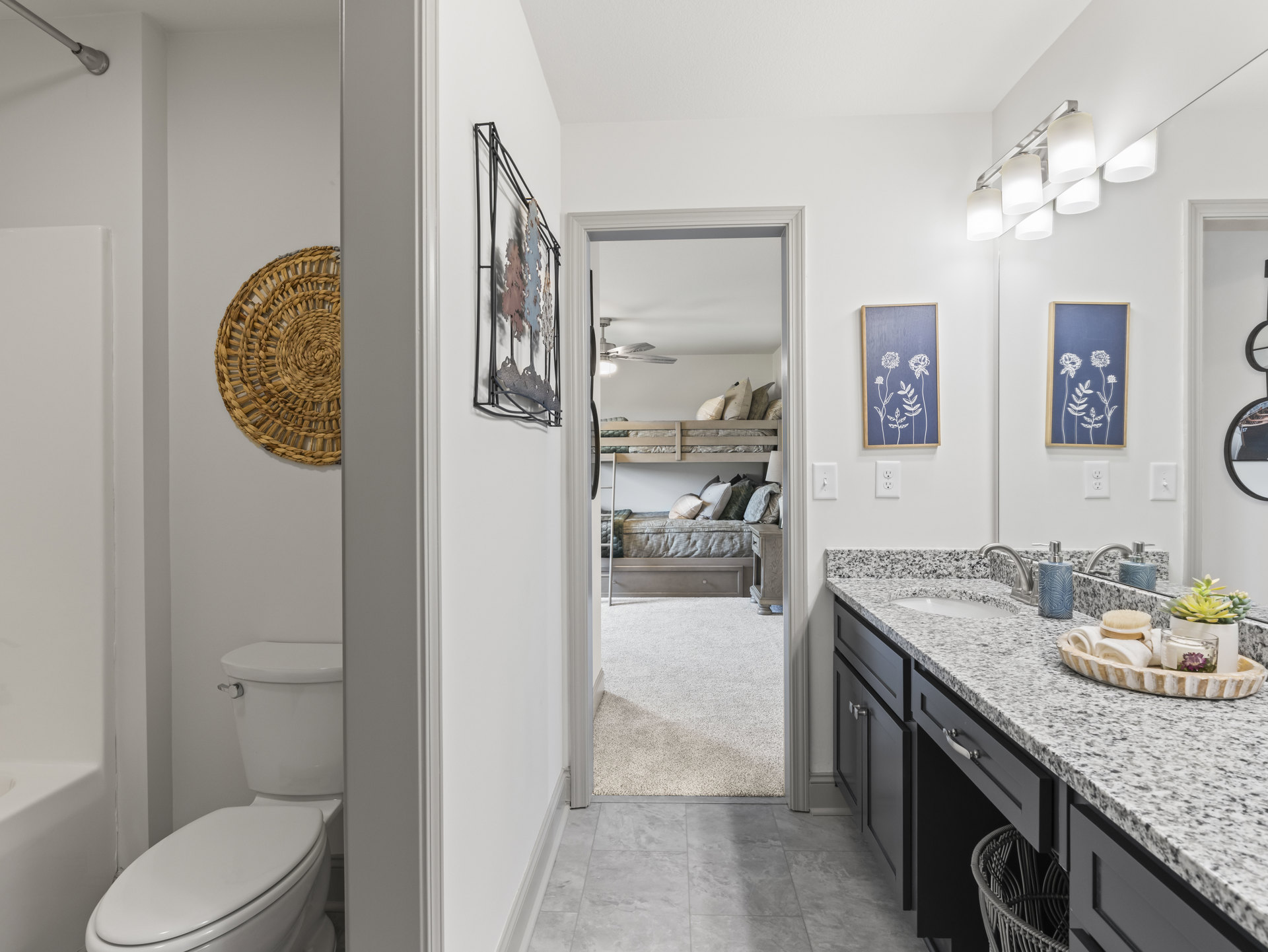 Bathroom with marble countertop sink, closed toilet seat, woven mat on tile floor, wall-mounted floral artwork, chalk drawing of flowers and leaves, large mirror above sink