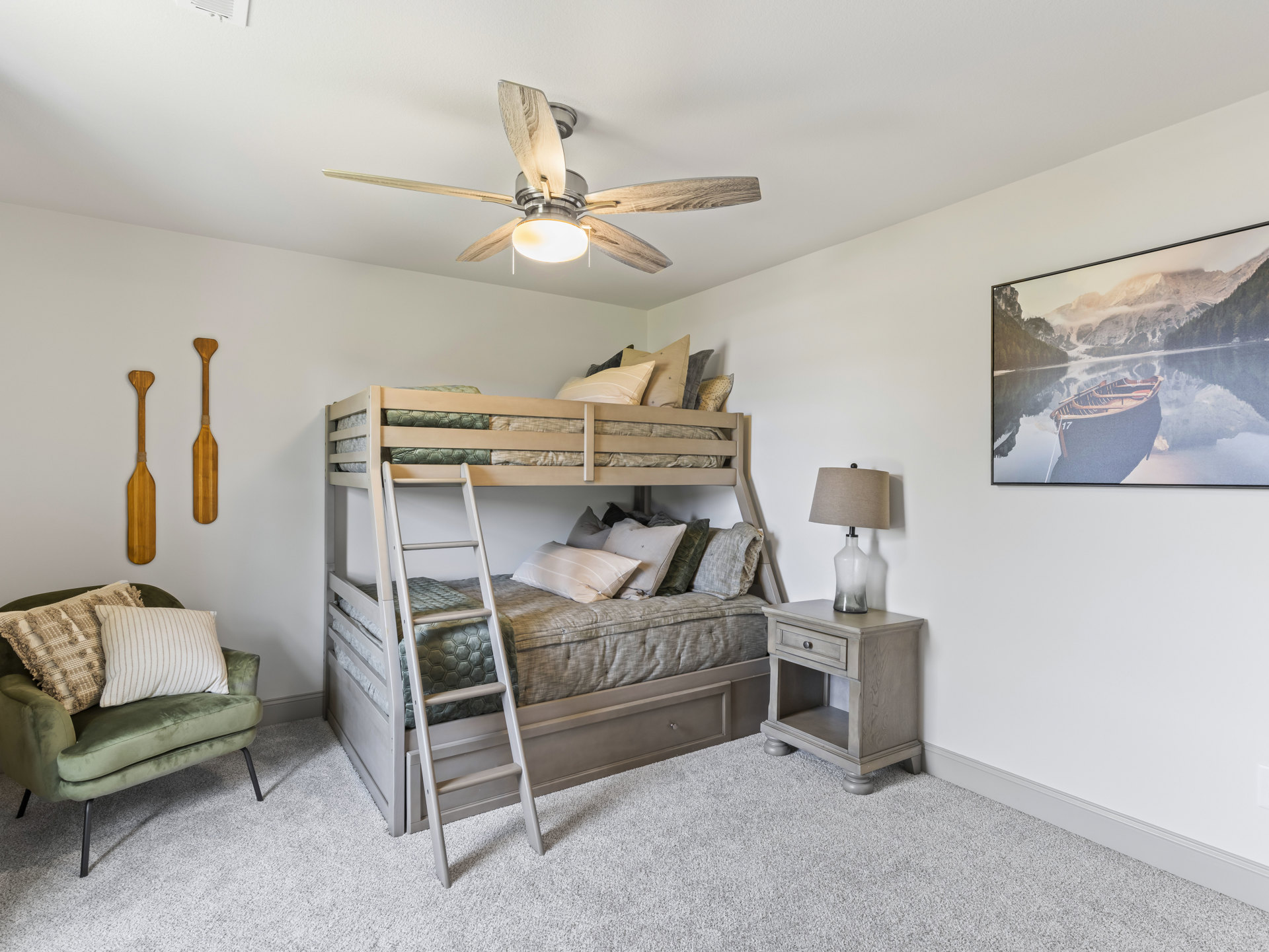 Wooden bunk bed with ladder, white bedding, framed boat picture on wall, ceiling fan with light, green accent chair with pillows, two pillows on gray couch