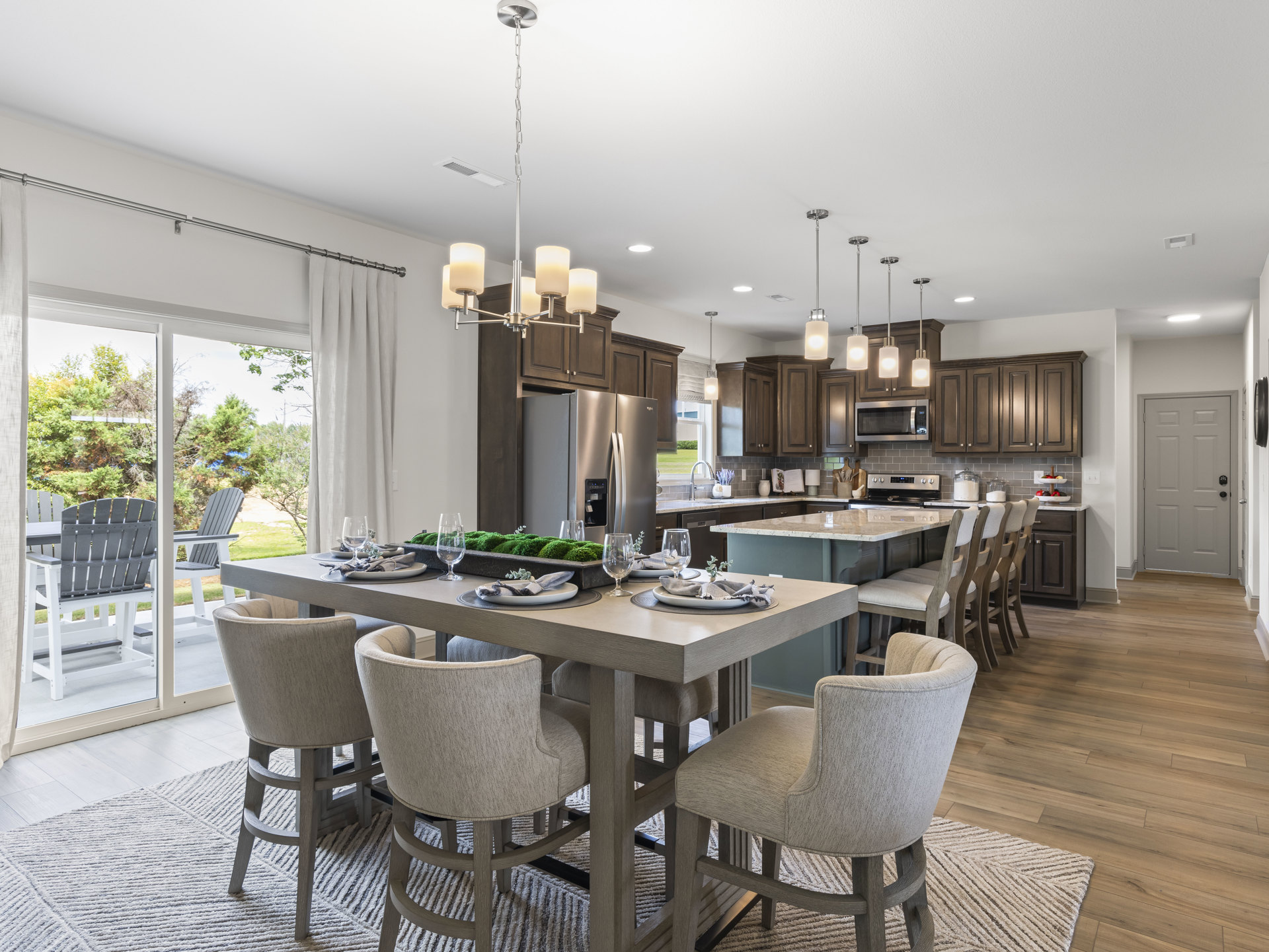 Open-concept kitchen and dining area featuring a wooden dining table set with plates and glasses, surrounded by upholstered chairs, light cabinetry, stainless steel microwave