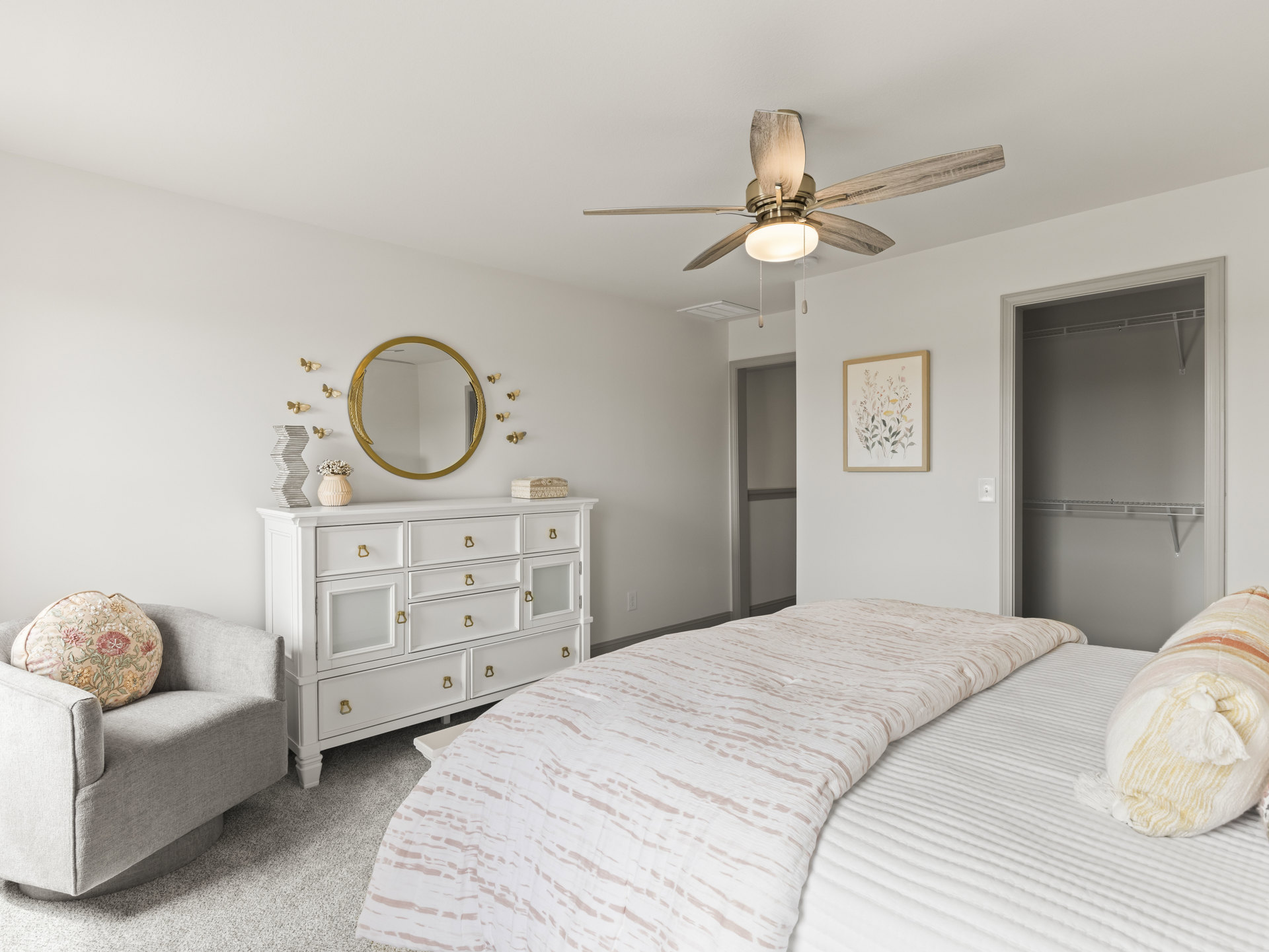 Bedroom with a white dresser topped by a round gold mirror, bed with white linens and blanket, floral painting in a frame on the wall, pillow on an upholstered chair, ceiling fan