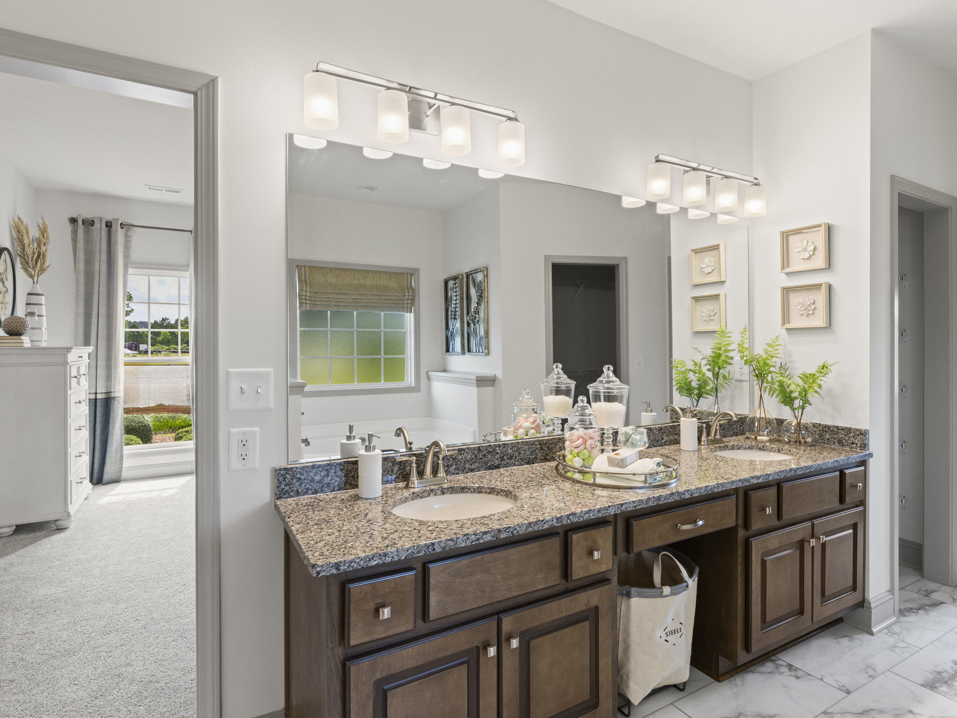 Bathroom with double sinks, wide frameless mirror, white quartz countertop, light wood cabinetry, chrome faucets, wall-mounted sconce lighting, decorative vase with dried plants