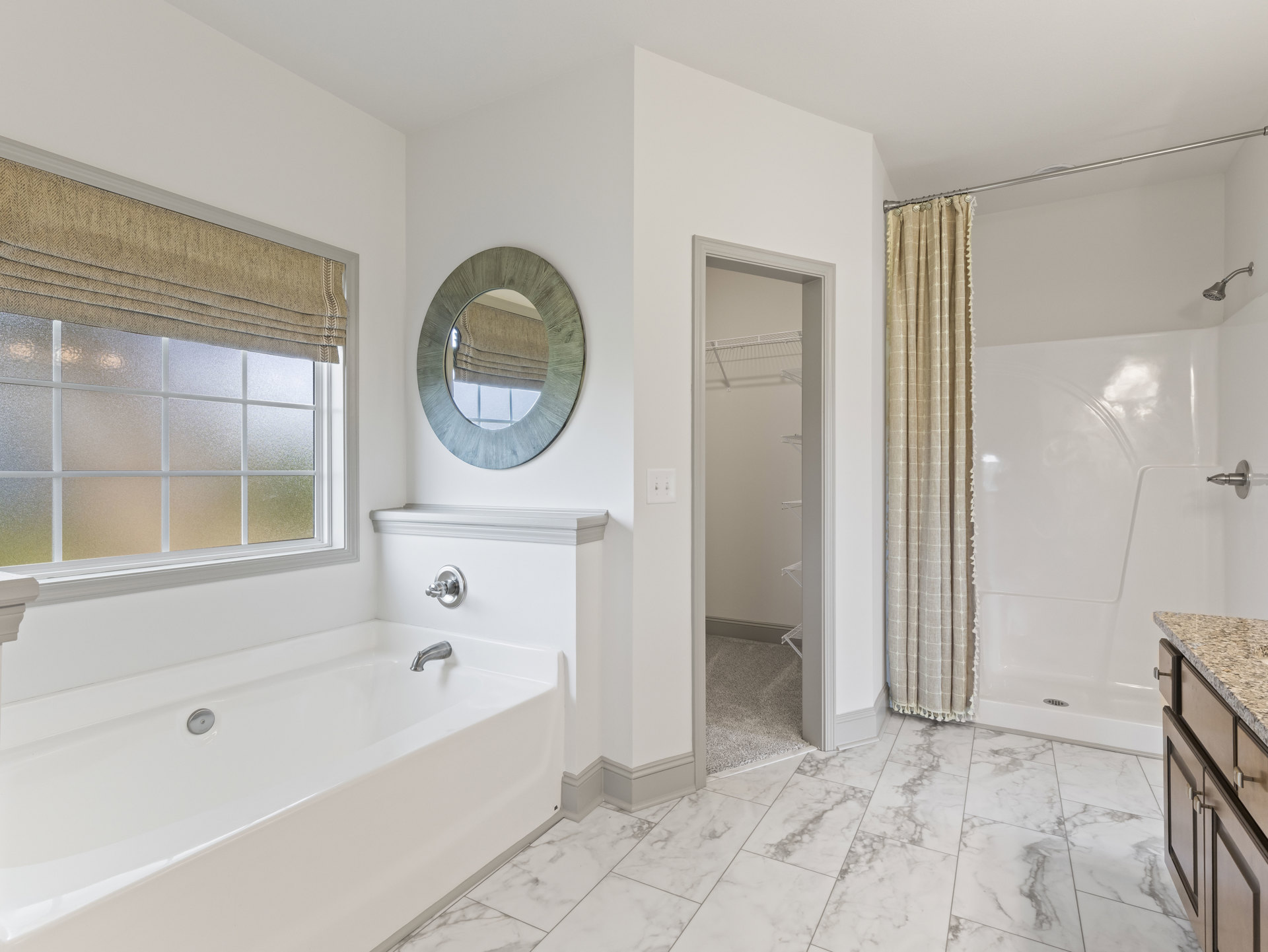White bathtub with silver faucet beside patterned shower curtain, wall-mounted mirror above sink, tiled walls, window with closed blind