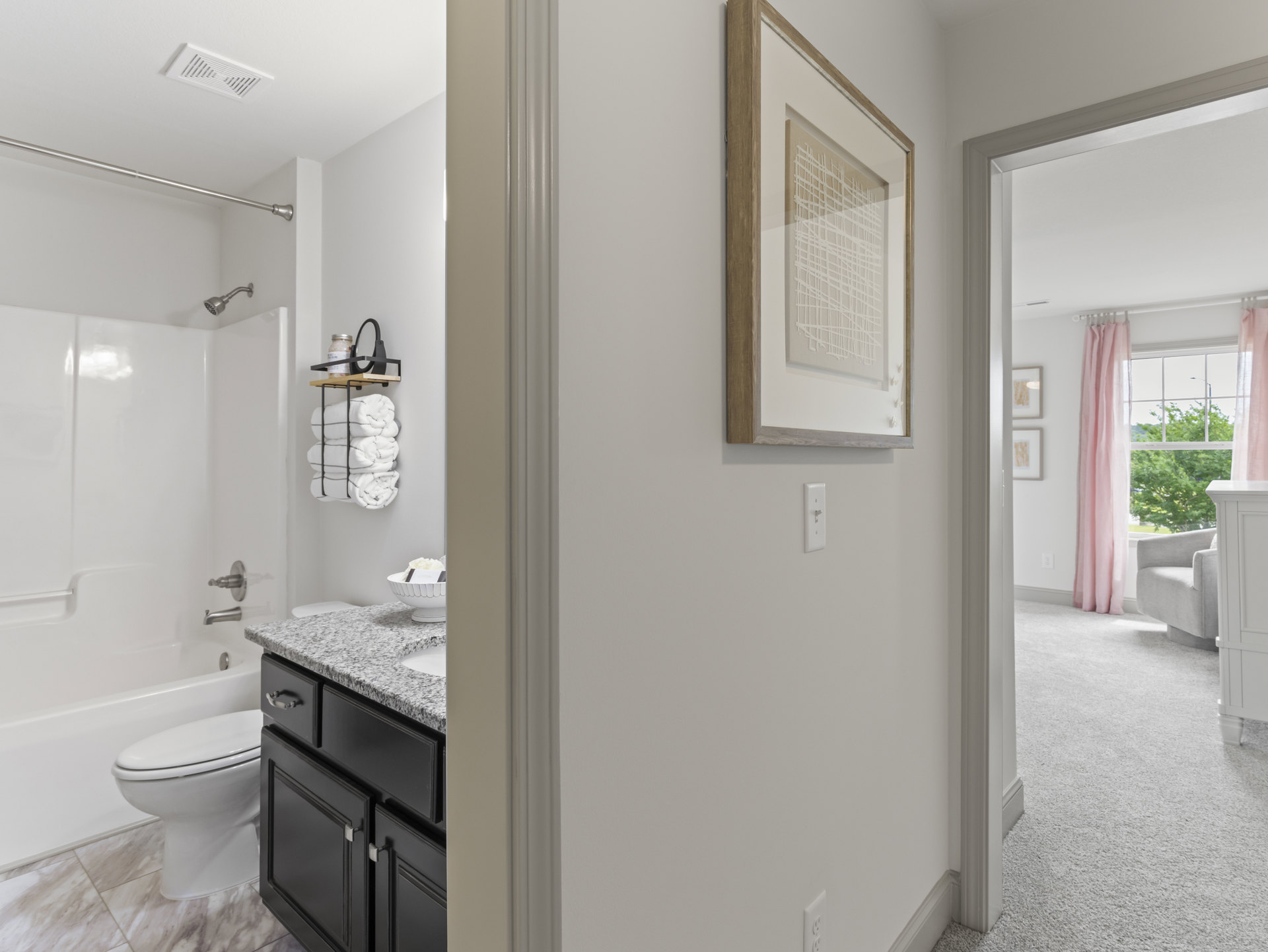 Bathroom with white toilet, rectangular sink on stone countertop, open shelving with folded towels, framed artwork on tiled wall, pink curtain, chrome faucet